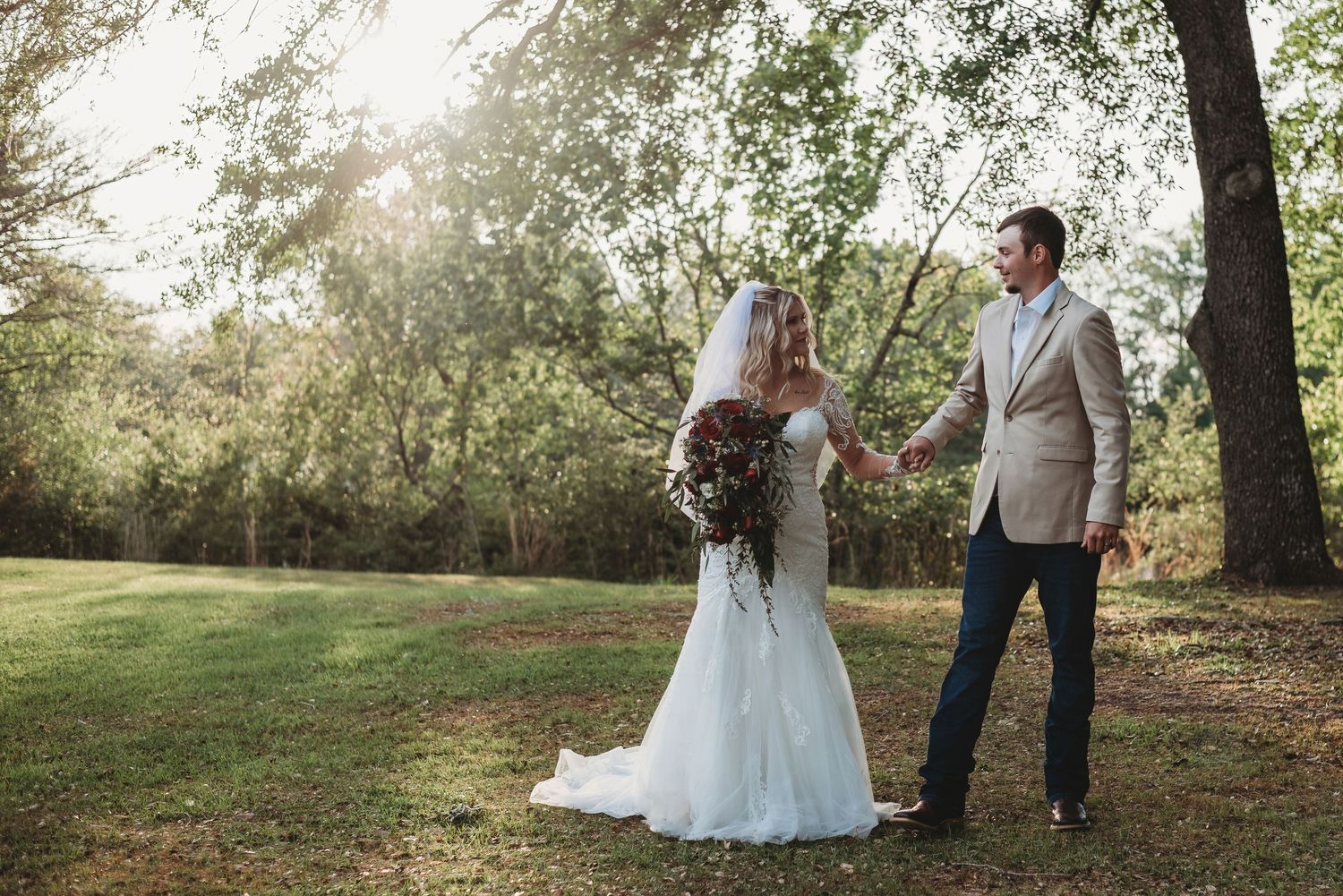 Bride and groom hold hands in a sunlit garden, she in a white gown with veil, he in a tan suit.