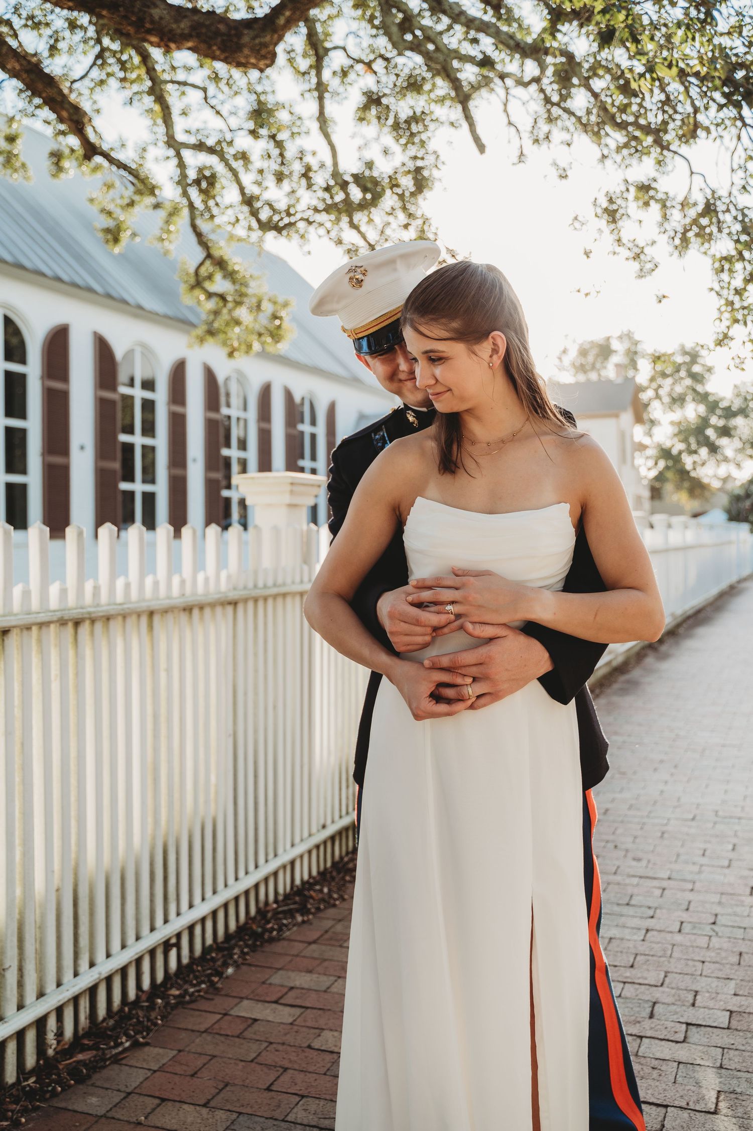 Marine in dress uniform embraces bride in white strapless gown near white picket fence at golden hour.