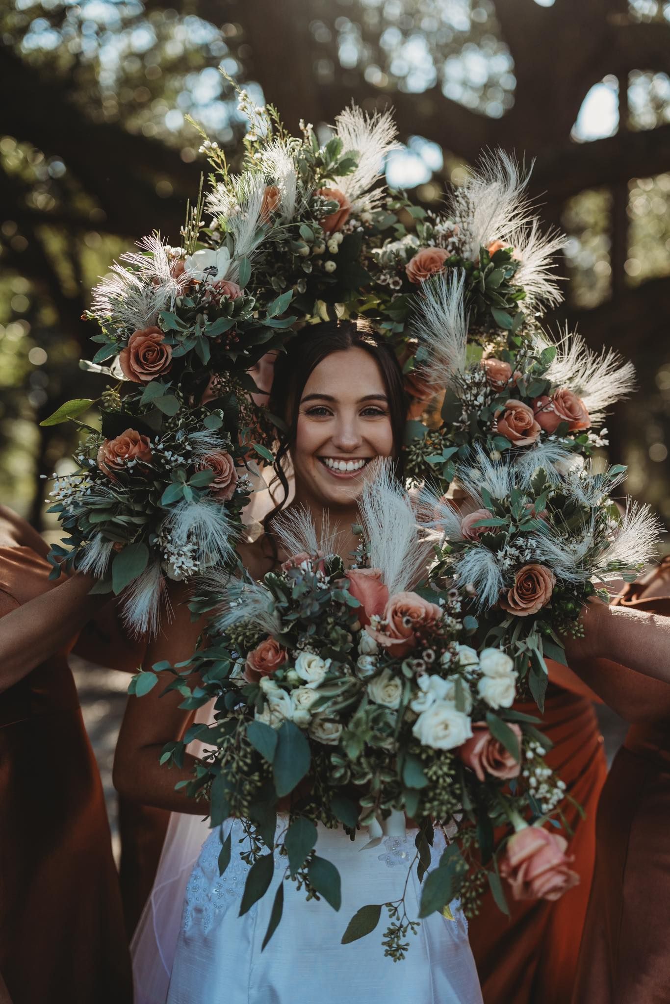 Joyful bride surrounded by large boho floral bouquets with roses, greenery, and feathers in an outdoor wedding setting.