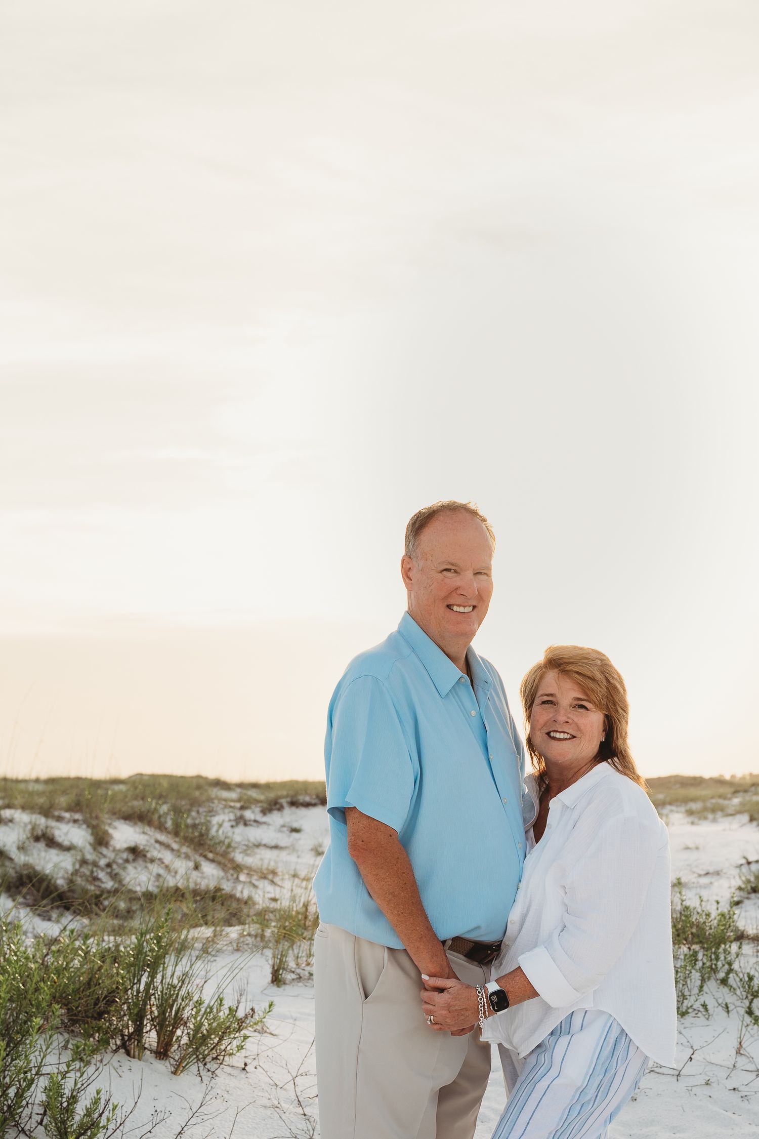 Smiling mature couple holding hands near sand dunes on a white sand beach at sunset.