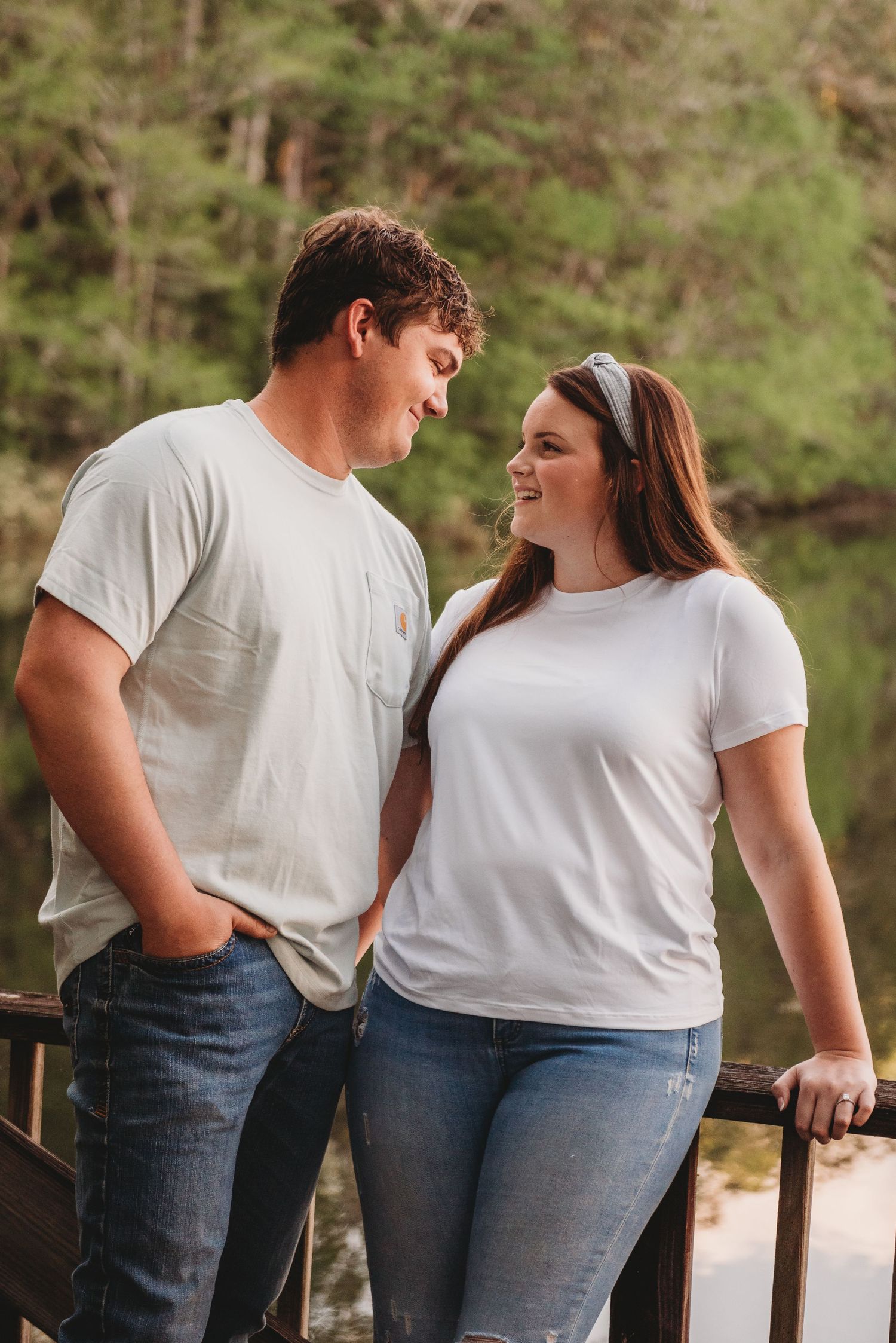 Happy couple smiling at each other while leaning on a wooden railing with lush green trees behind.