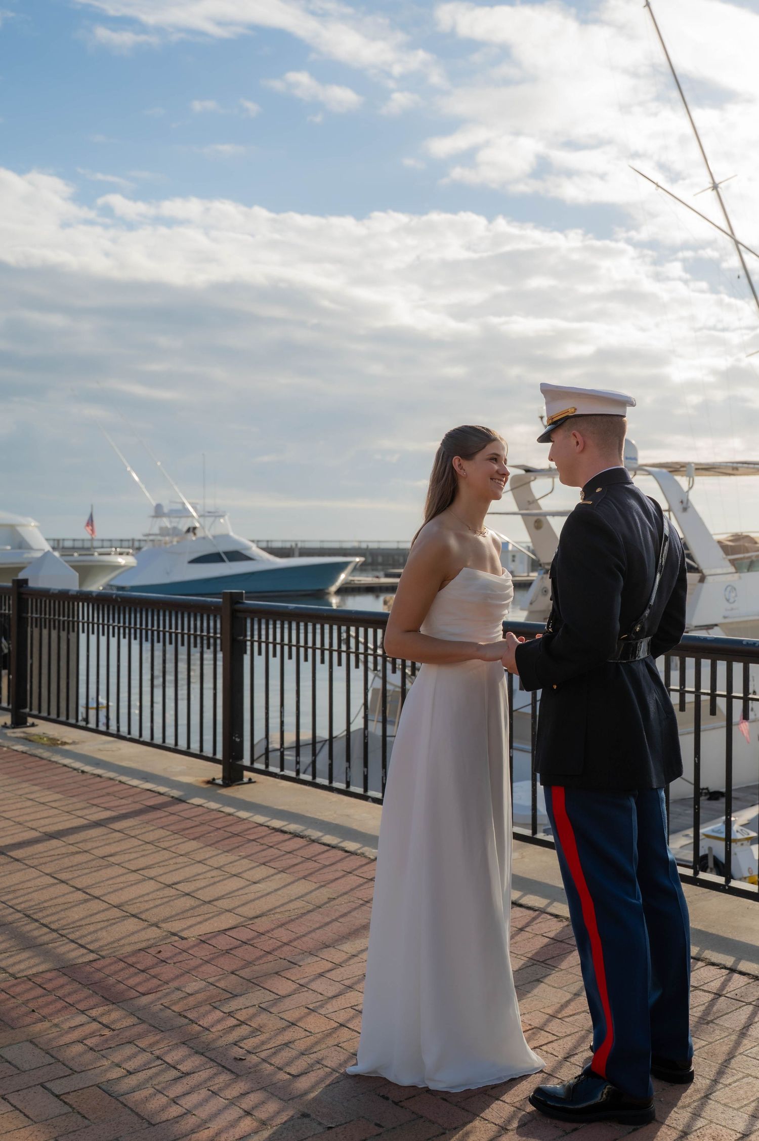 Marine in dress uniform holds hands with bride in white gown on waterfront boardwalk with yachts in background.