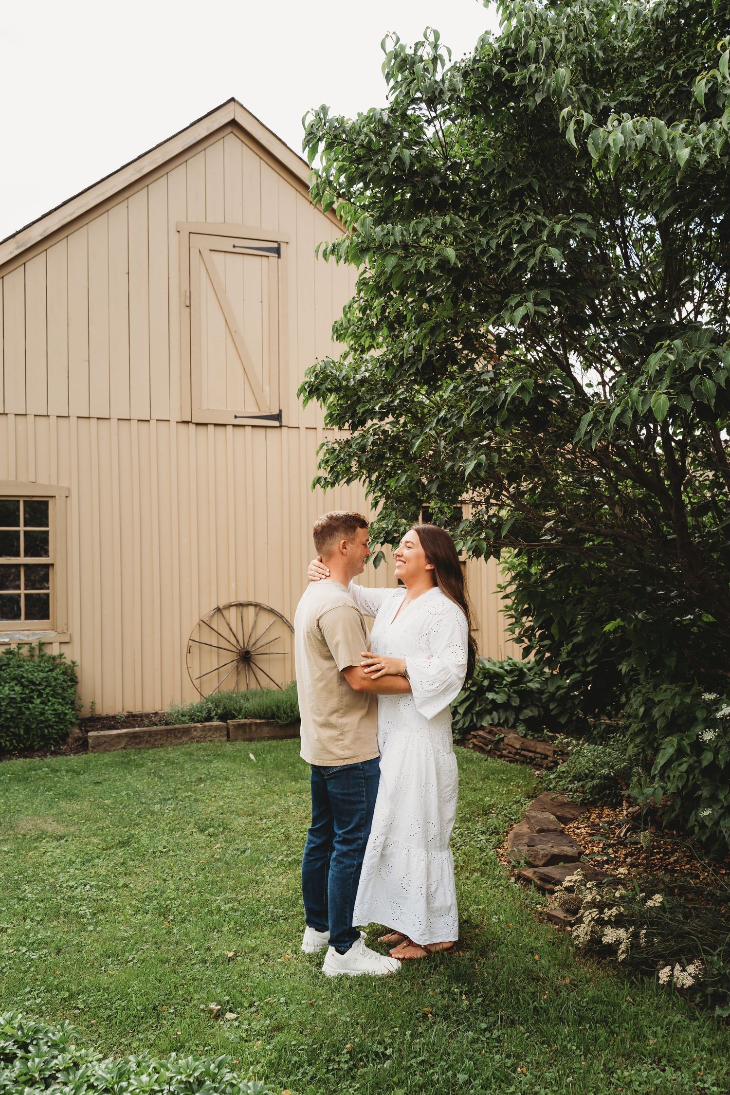 Couple embracing in front of a rustic beige barn with a wagon wheel, surrounded by lush green trees and grass.