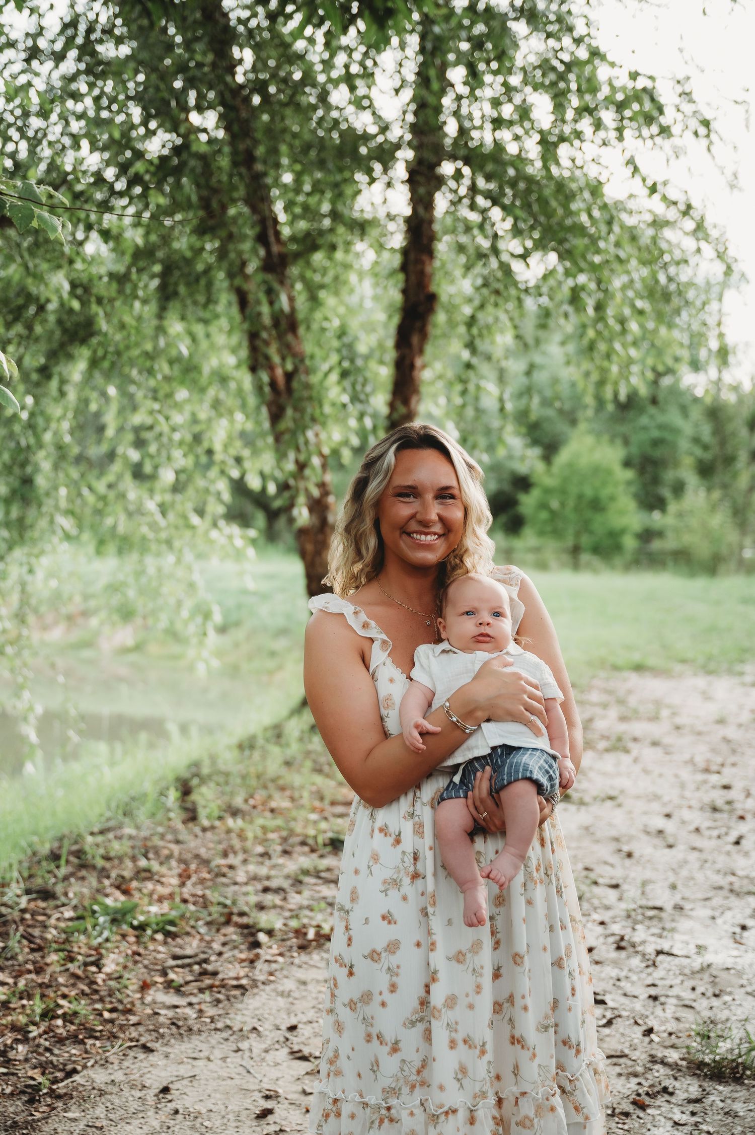 Smiling woman in floral dress holding a baby outdoors near a willow tree on a dirt path.
