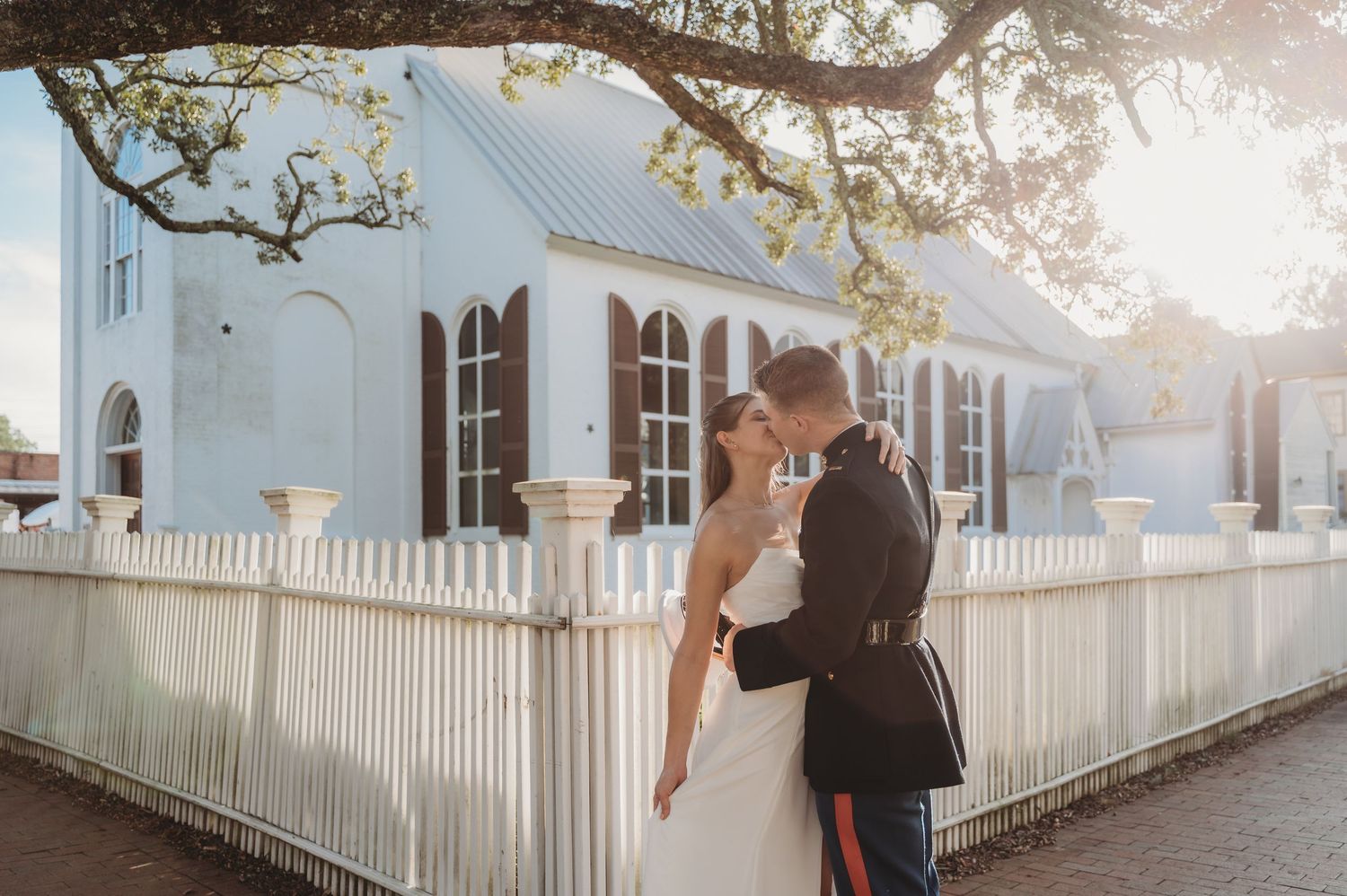 Military groom in dress uniform kissing bride in white gown outside historic white church with picket fence.
