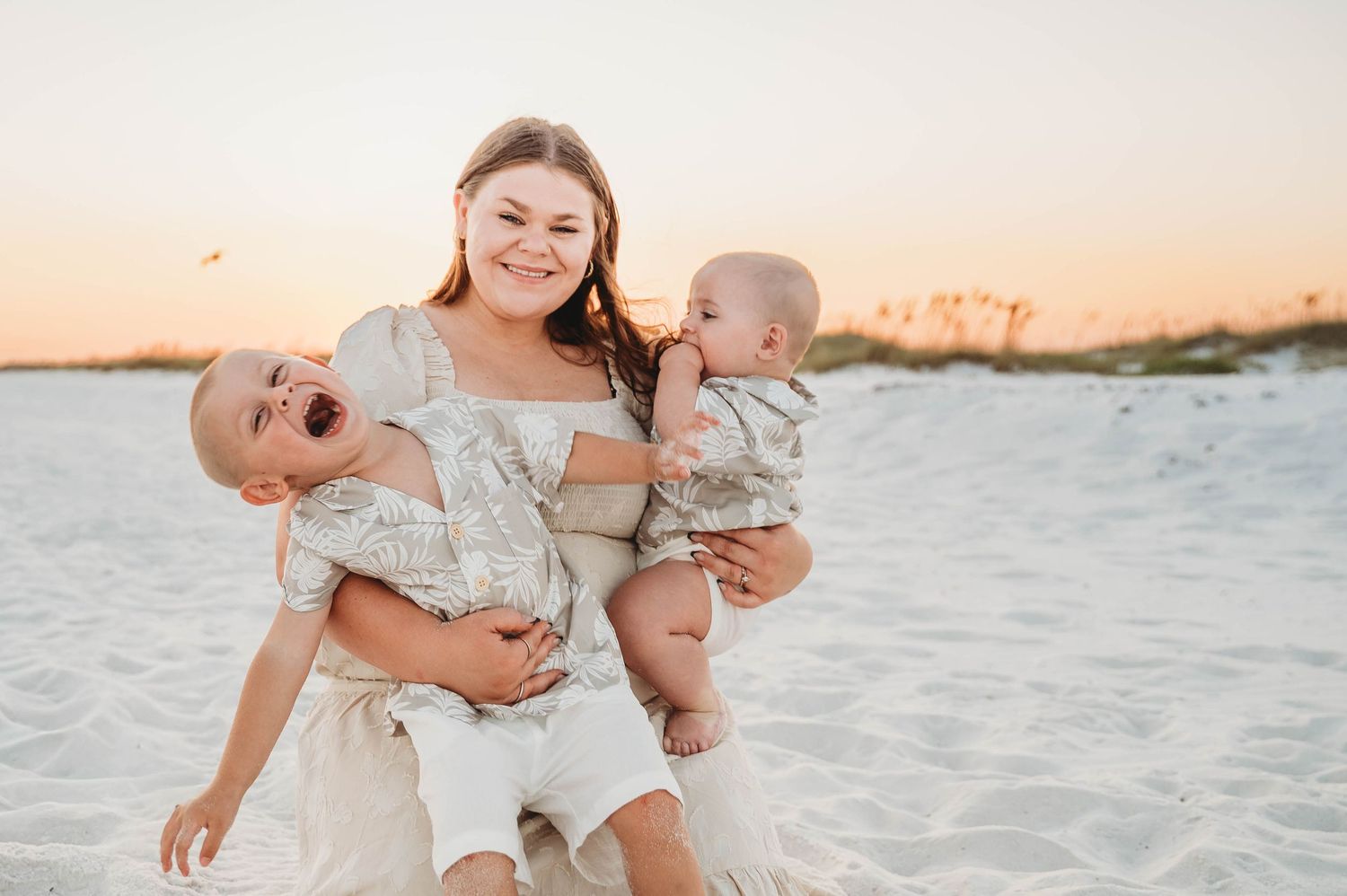 A smiling mother holds two babies on a sandy beach at sunset.
