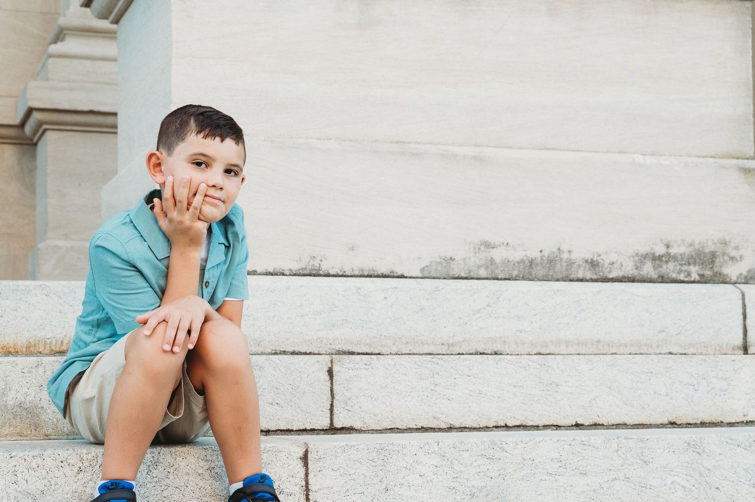 A young boy in a blue shirt and shorts sits thoughtfully on stone steps outdoors.