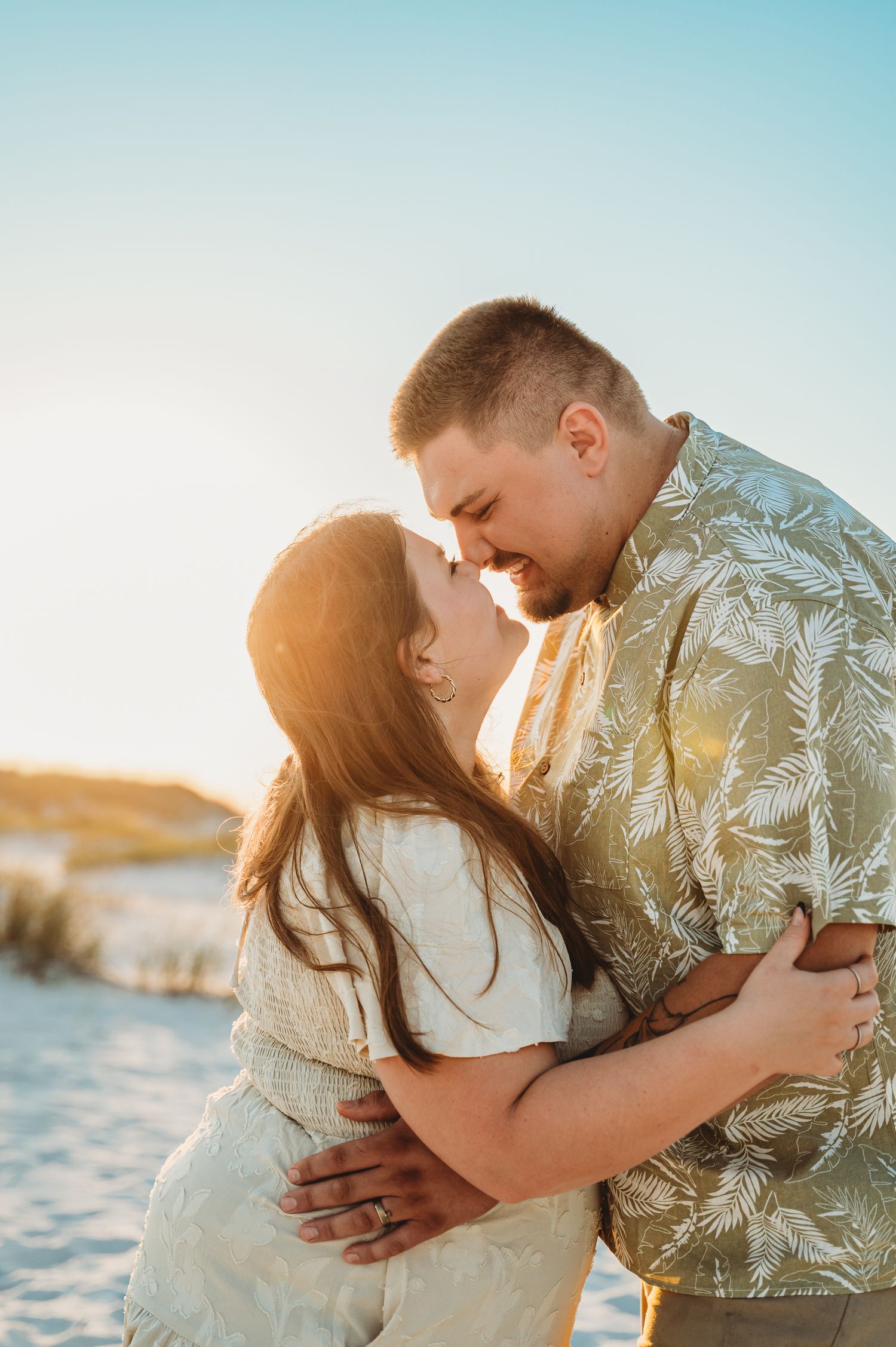 Couple nearly kissing on a sunny beach with golden backlight and sand dunes.