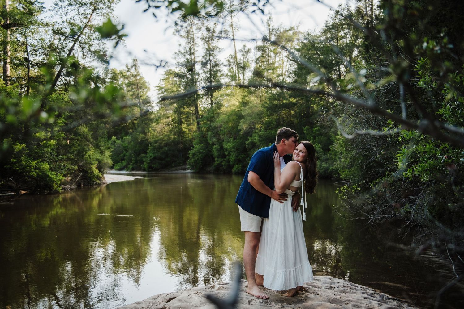 Couple sharing a kiss on rocky riverbank framed by green foliage and calm reflective water.