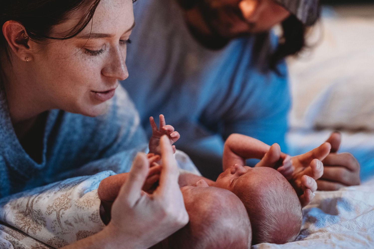 Parents lying beside newborn baby on bed, gently holding tiny hands and feet.