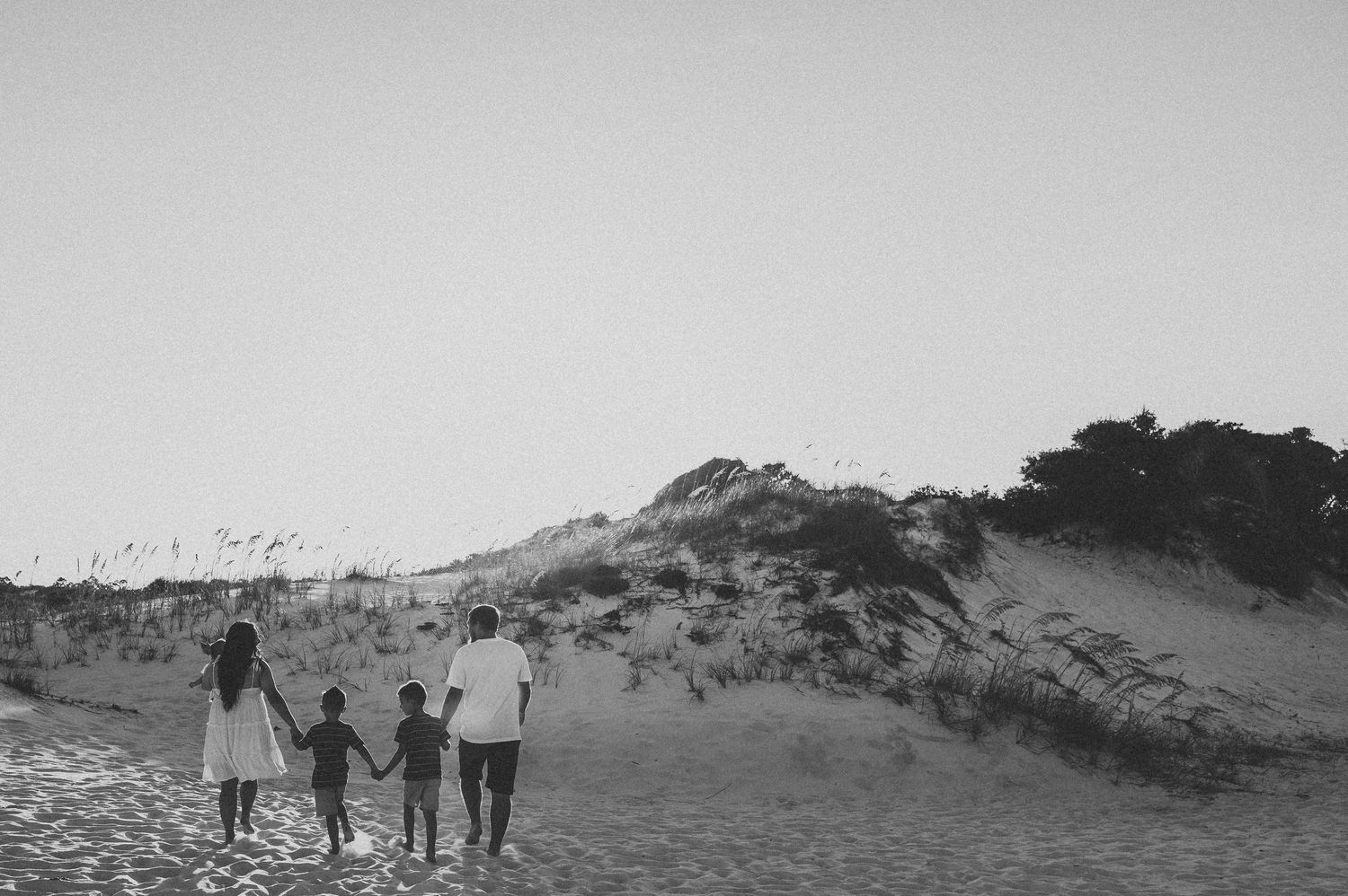 Black and white photo of family holding hands walking toward sandy beach dunes.