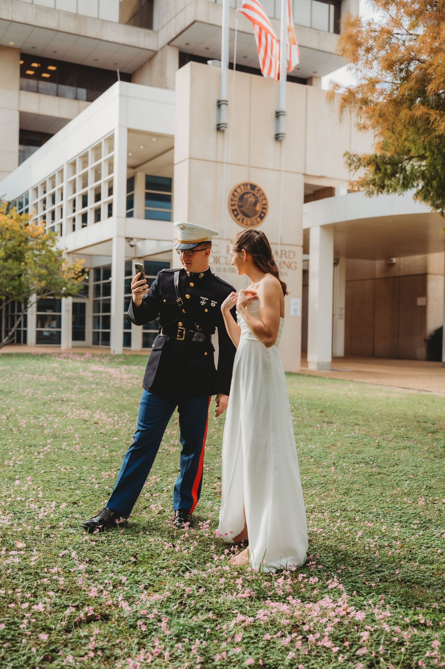 Marine groom shows bride something on phone outside government building on wedding day.
