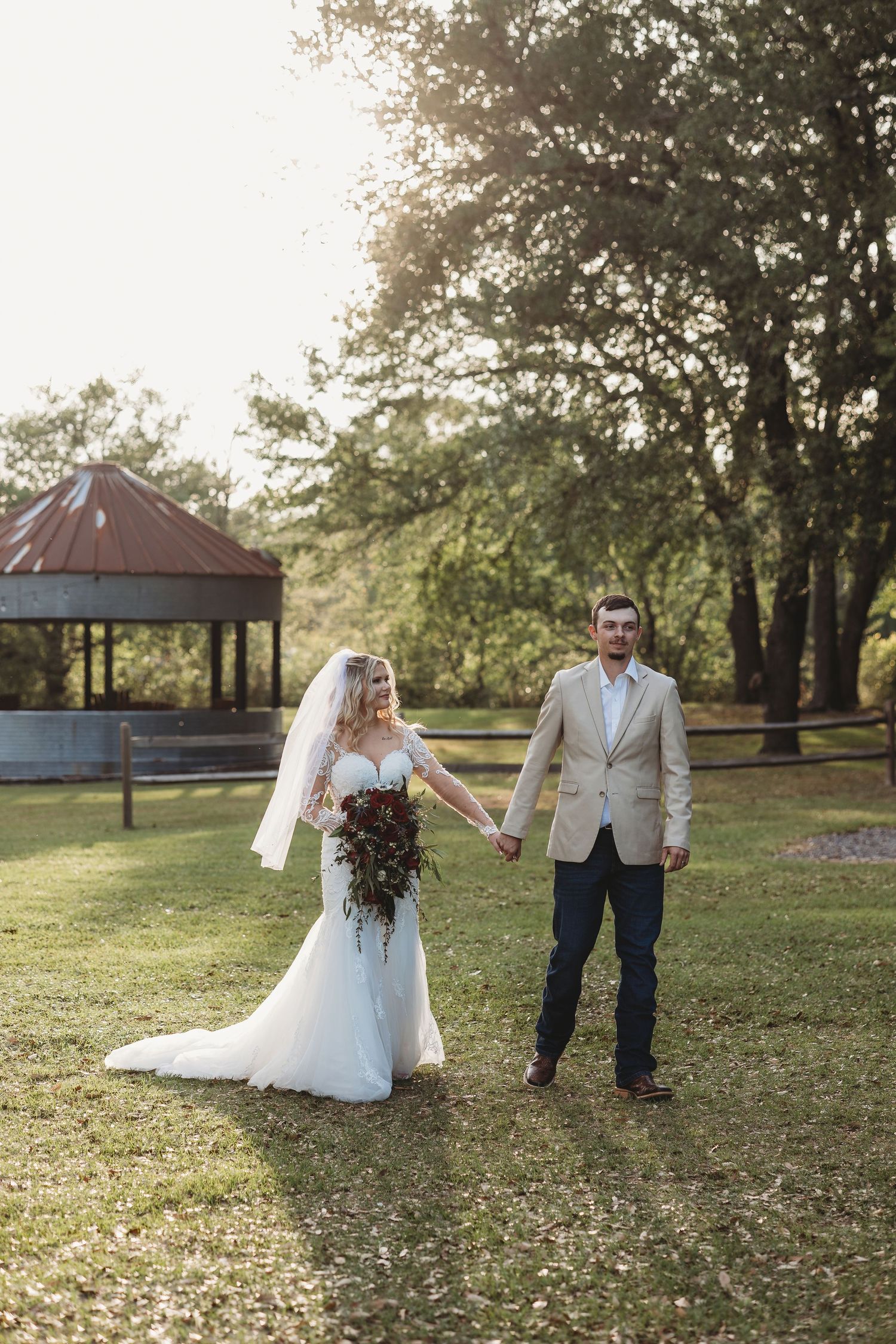 Bride in white gown holding bouquet and groom in tan blazer hold hands outdoors on green lawn at golden hour.
