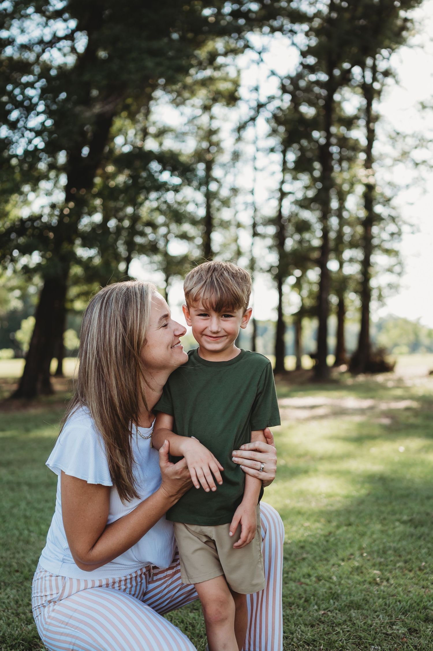 A woman crouches behind a smiling boy in a green shirt outdoors, surrounded by tall trees in a sunny park.