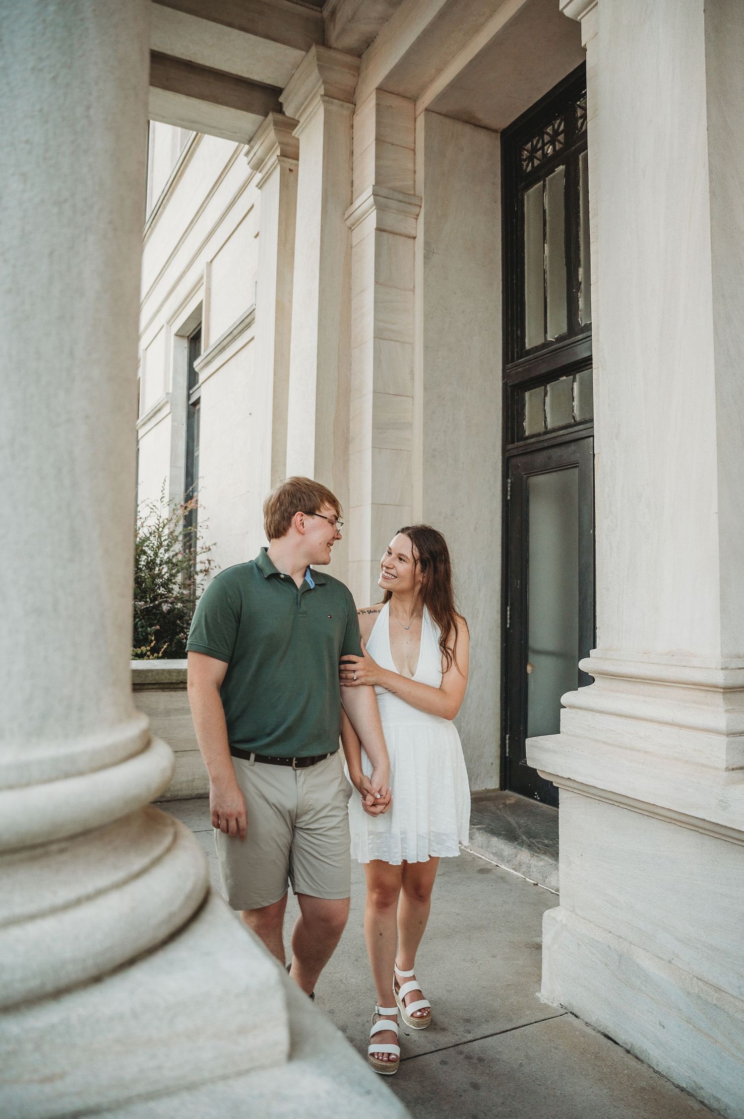 Couple walking hand in hand past white columns of a classic building, smiling at each other.