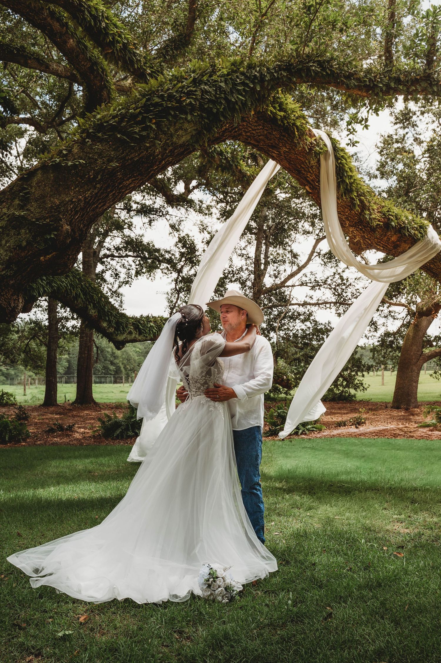 Bride and groom share a romantic moment under a moss-covered oak tree with white draping.