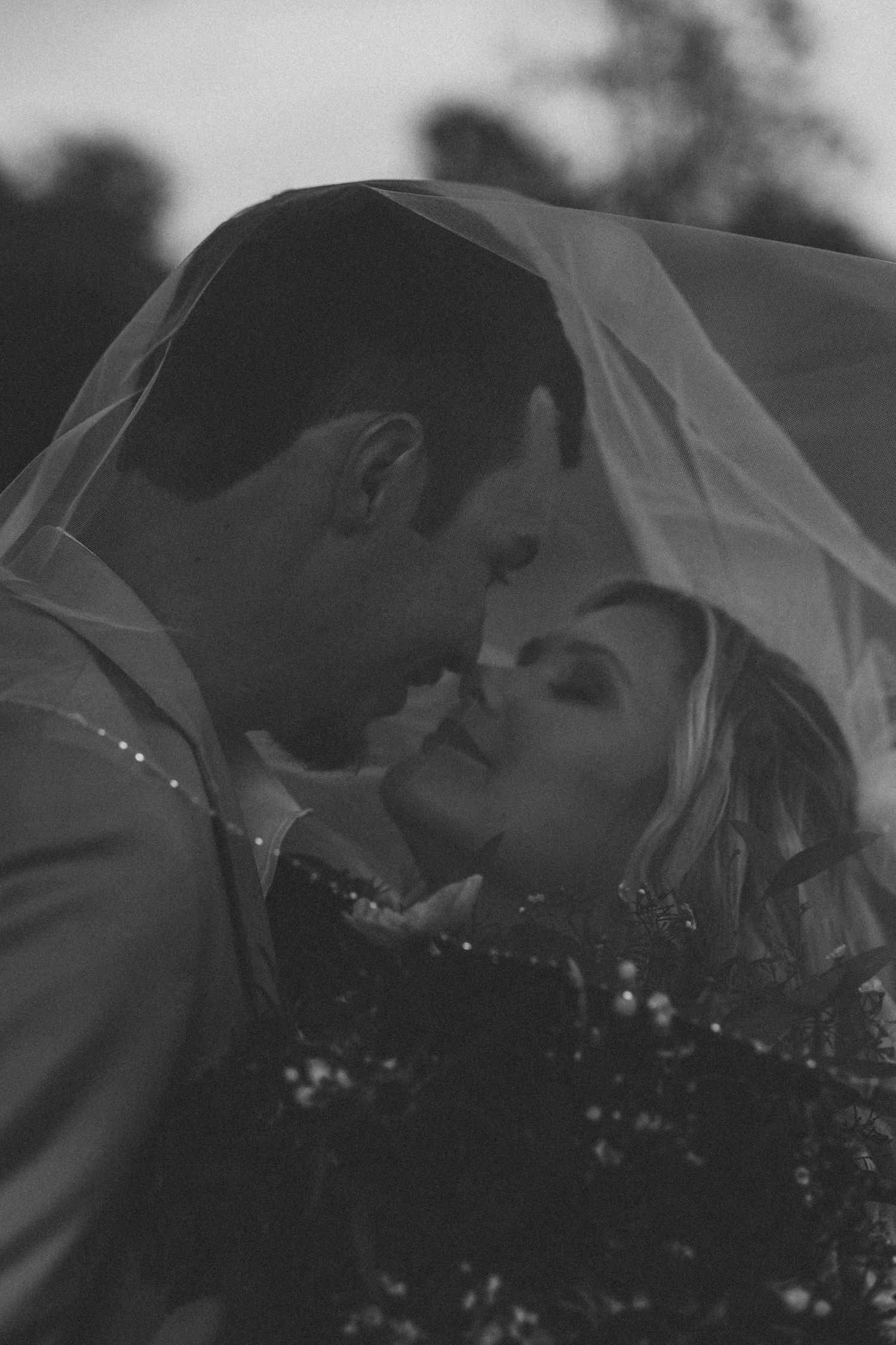 Black and white romantic photo of bride and groom nearly kissing beneath a flowing veil.