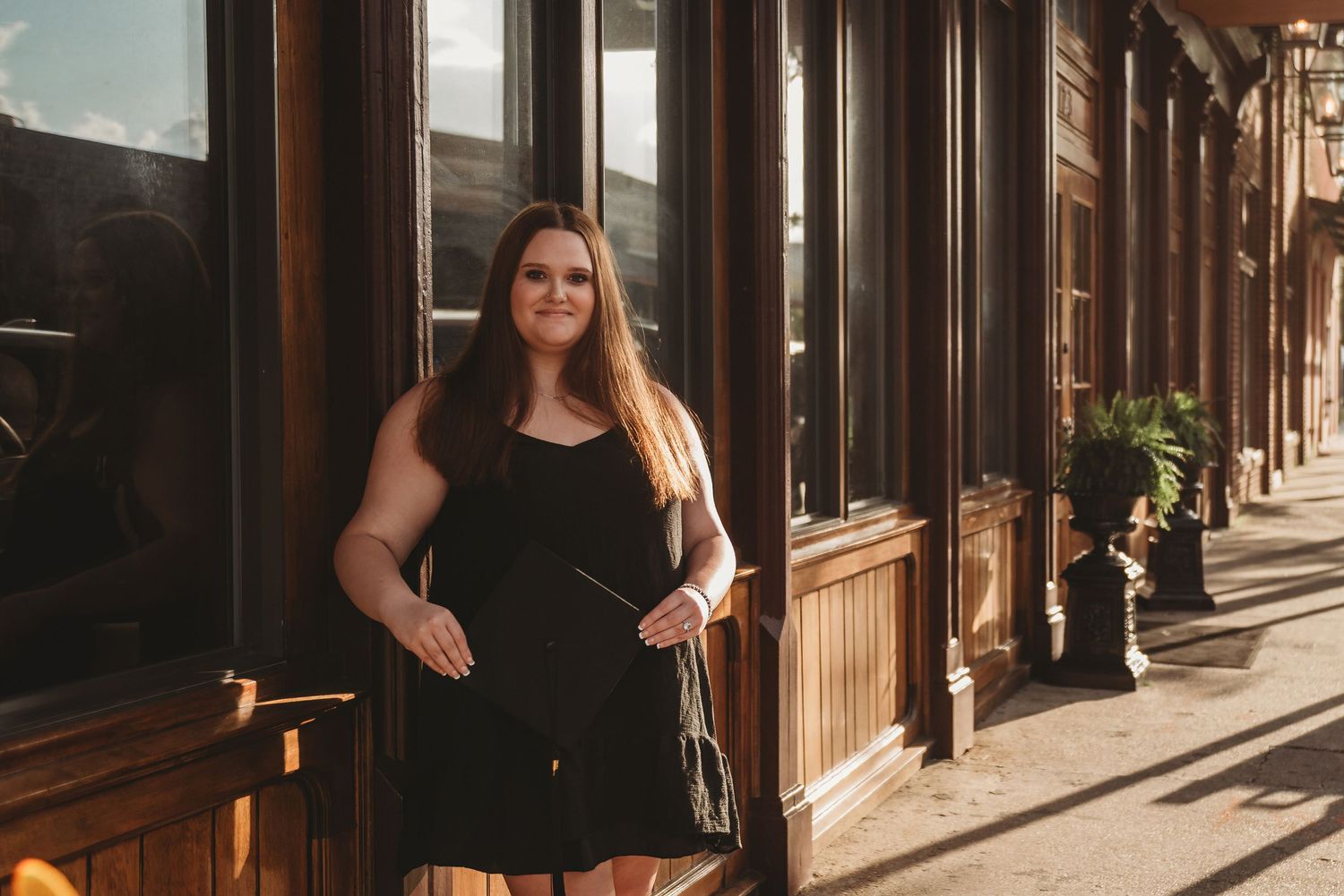 A woman in a black dress leans against a storefront in golden sunlight.