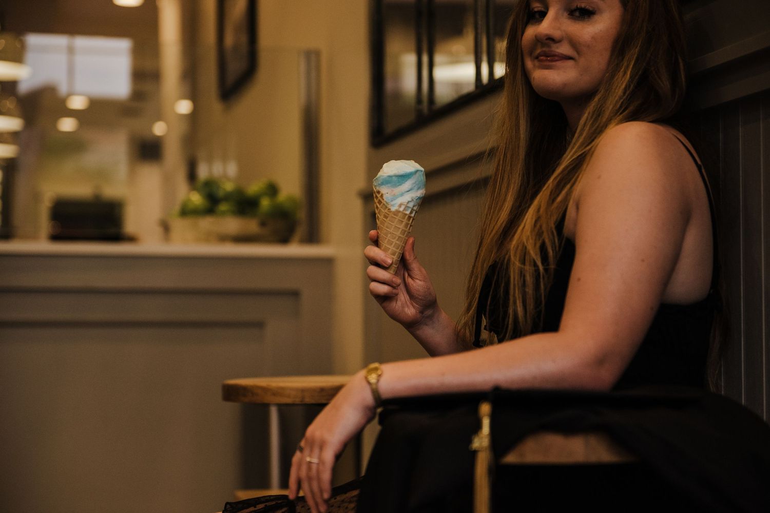 Young woman in black dress holding a blue ice cream cone while sitting in a chair at an ice cream shop.