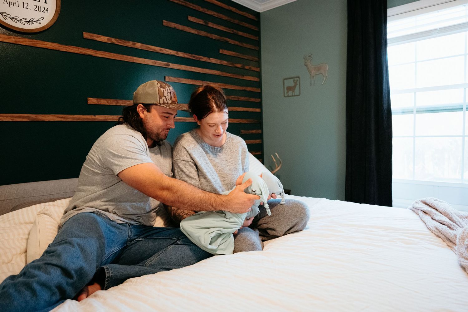 New parents sitting on a white bed together lovingly looking at their swaddled newborn baby.