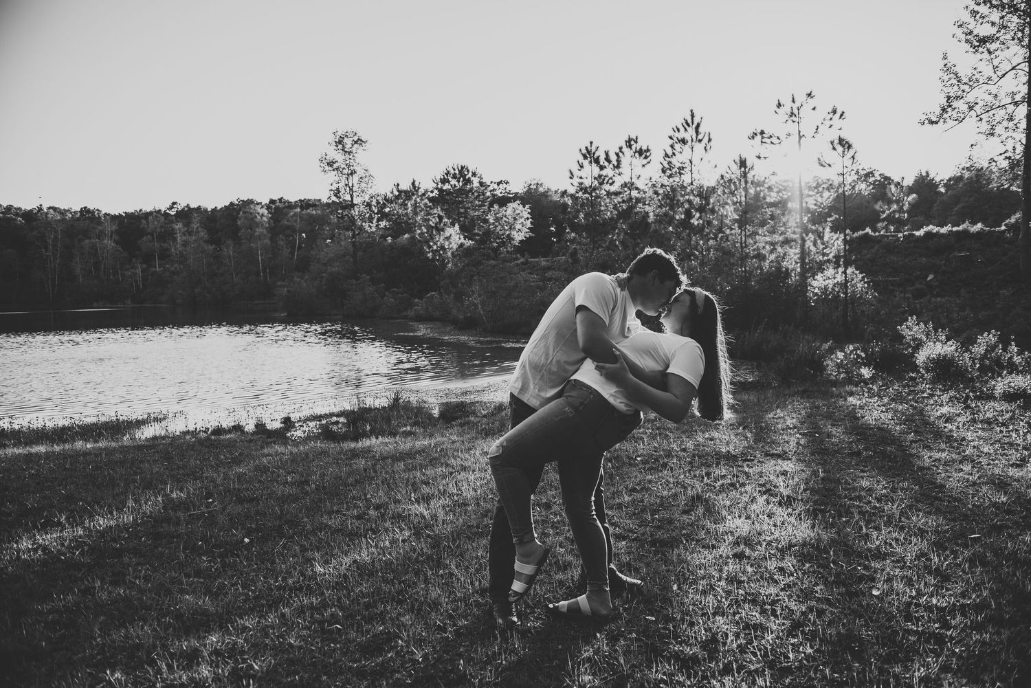 Black and white romantic photo of couple sharing a dip kiss near a lake in natural surroundings.