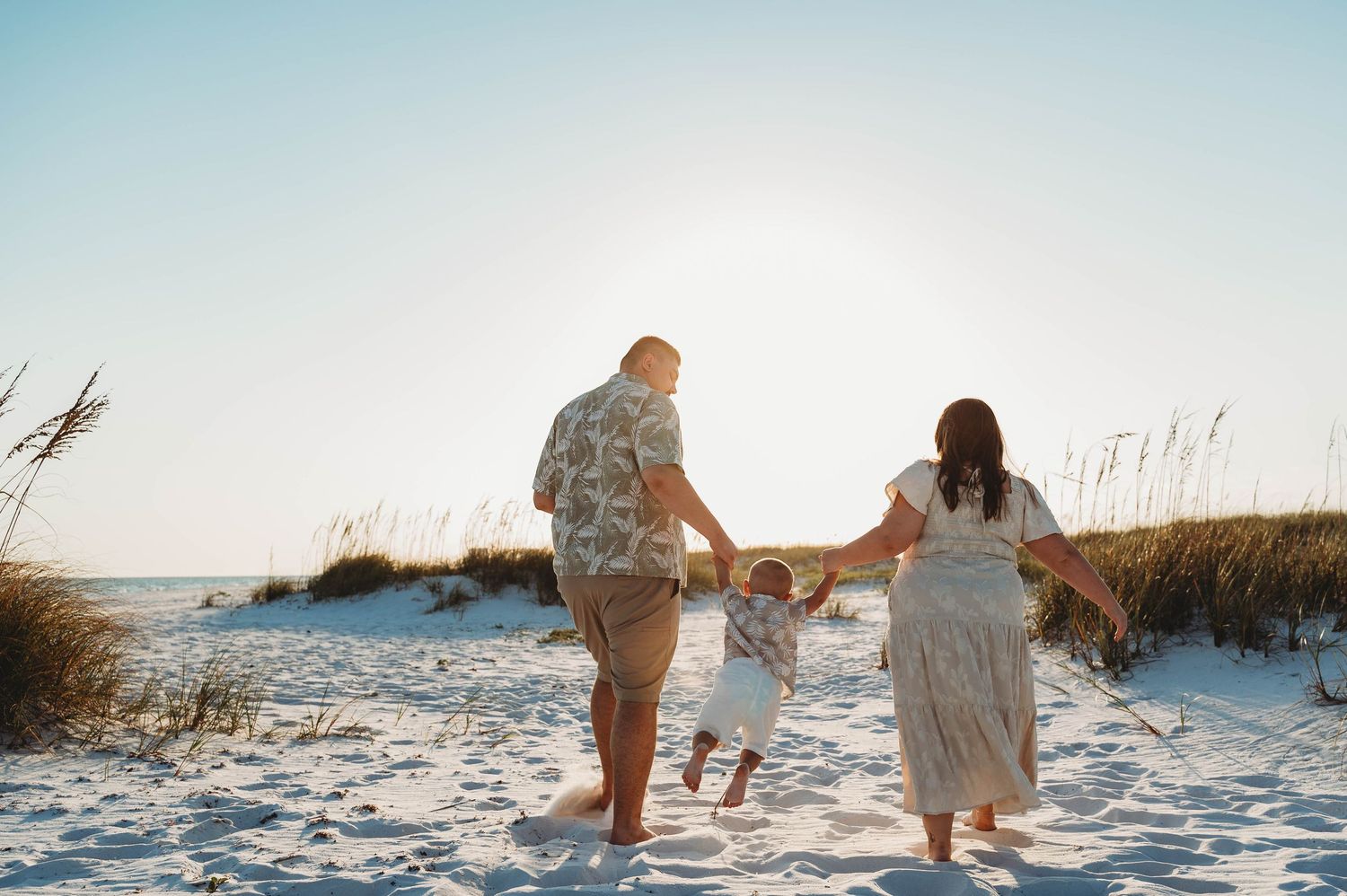 Family walking toward ocean on white sandy beach holding toddlers hands.