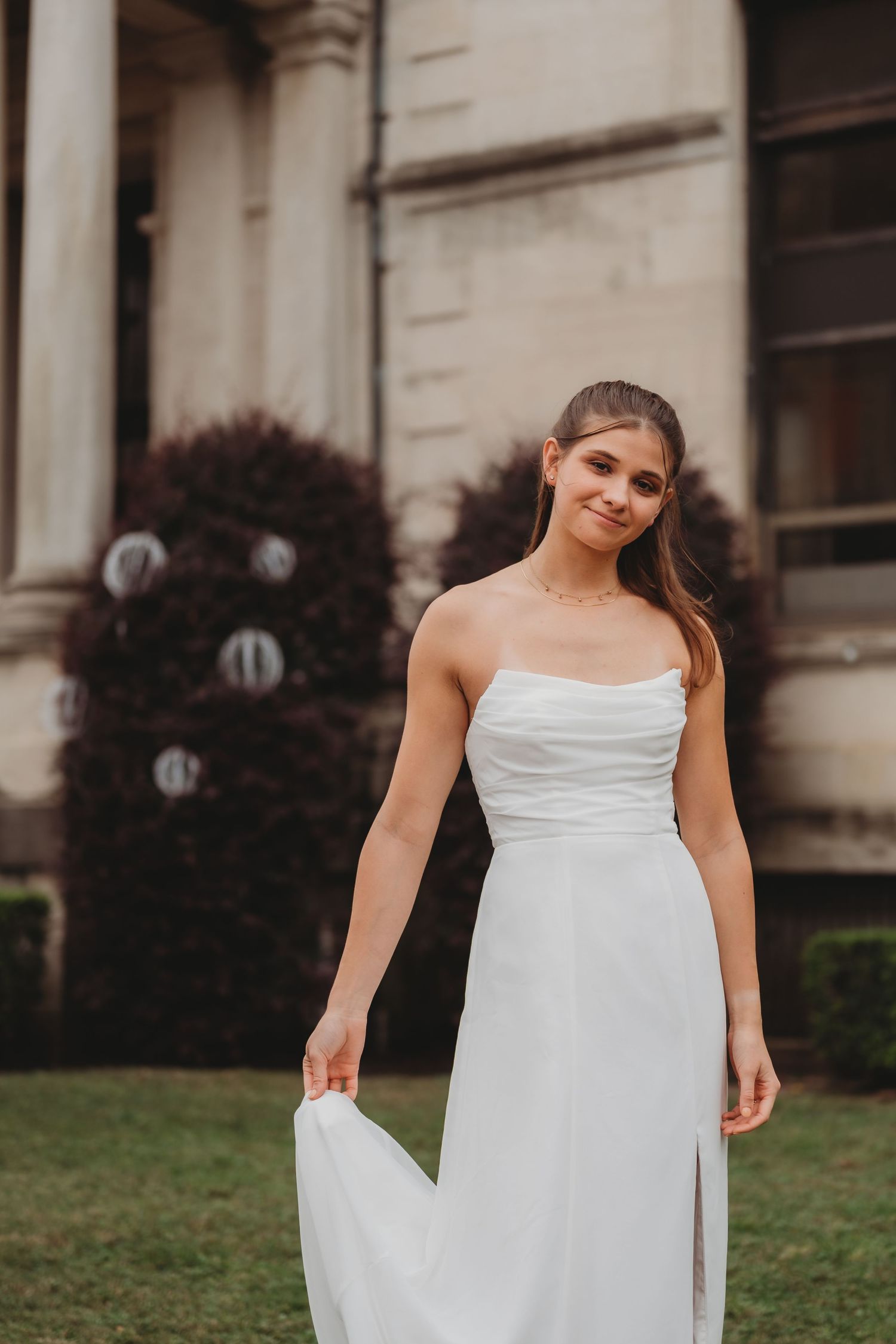 Young woman in a white strapless wedding gown smiling outdoors near a classic building.