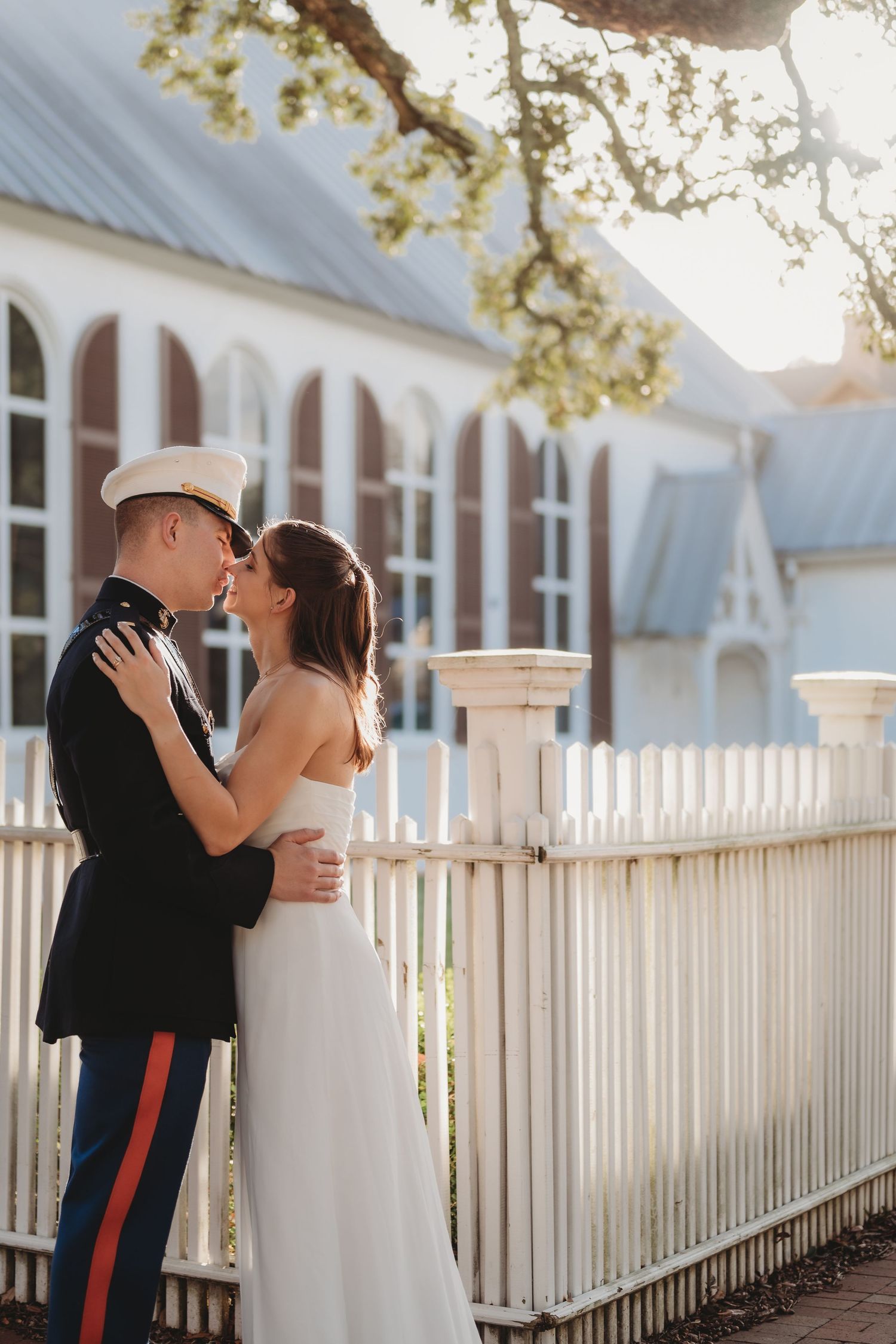 Marine in dress uniform embraces bride in white gown by white picket fence in golden hour light.