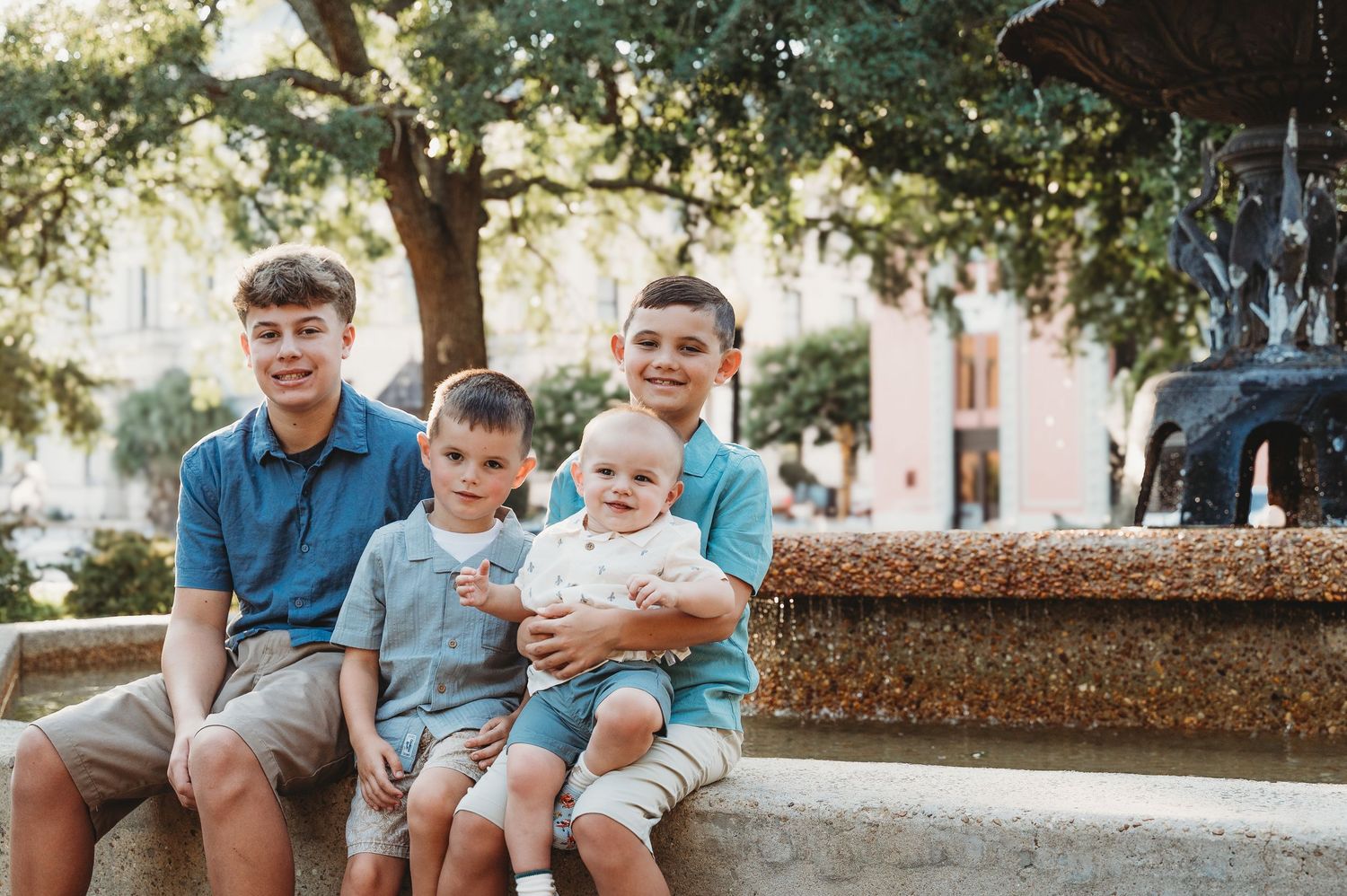 Four brothers sit together by a decorative fountain, including a smiling baby, in an outdoor plaza.