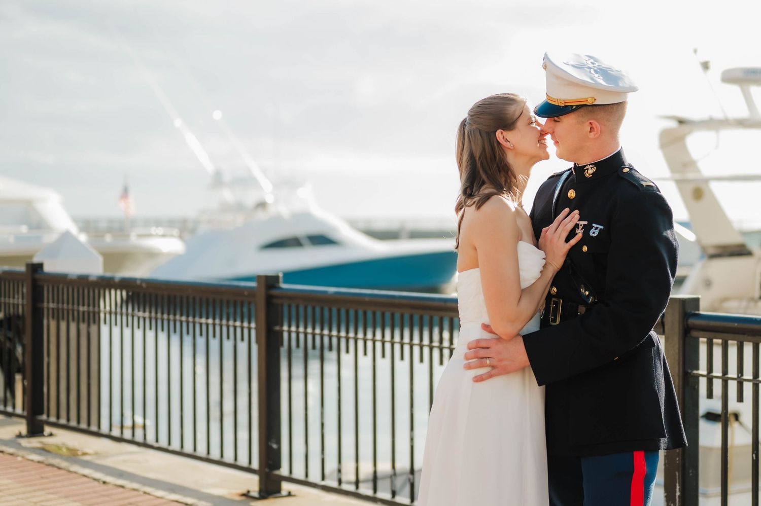 Marine in dress uniform shares a romantic kiss with bride on a waterfront dock with yachts in the background.