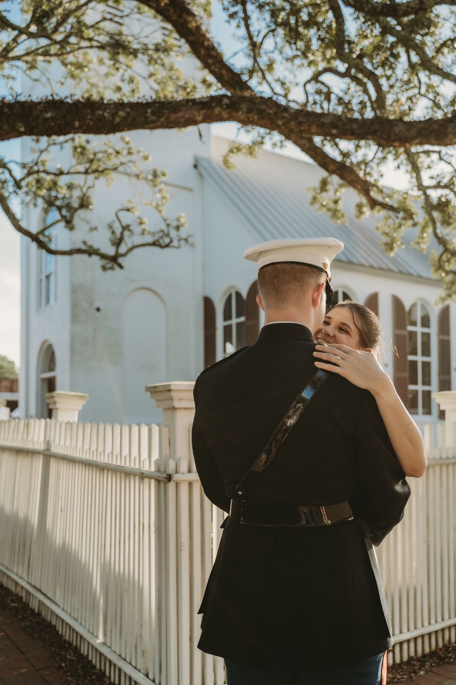 Marine groom embraces smiling bride near white fence and blue historic church.