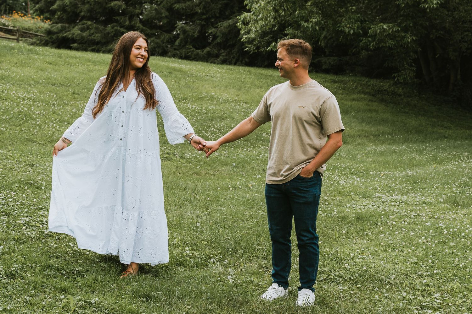 A couple holds hands and smiles while walking on a green grassy field surrounded by trees.