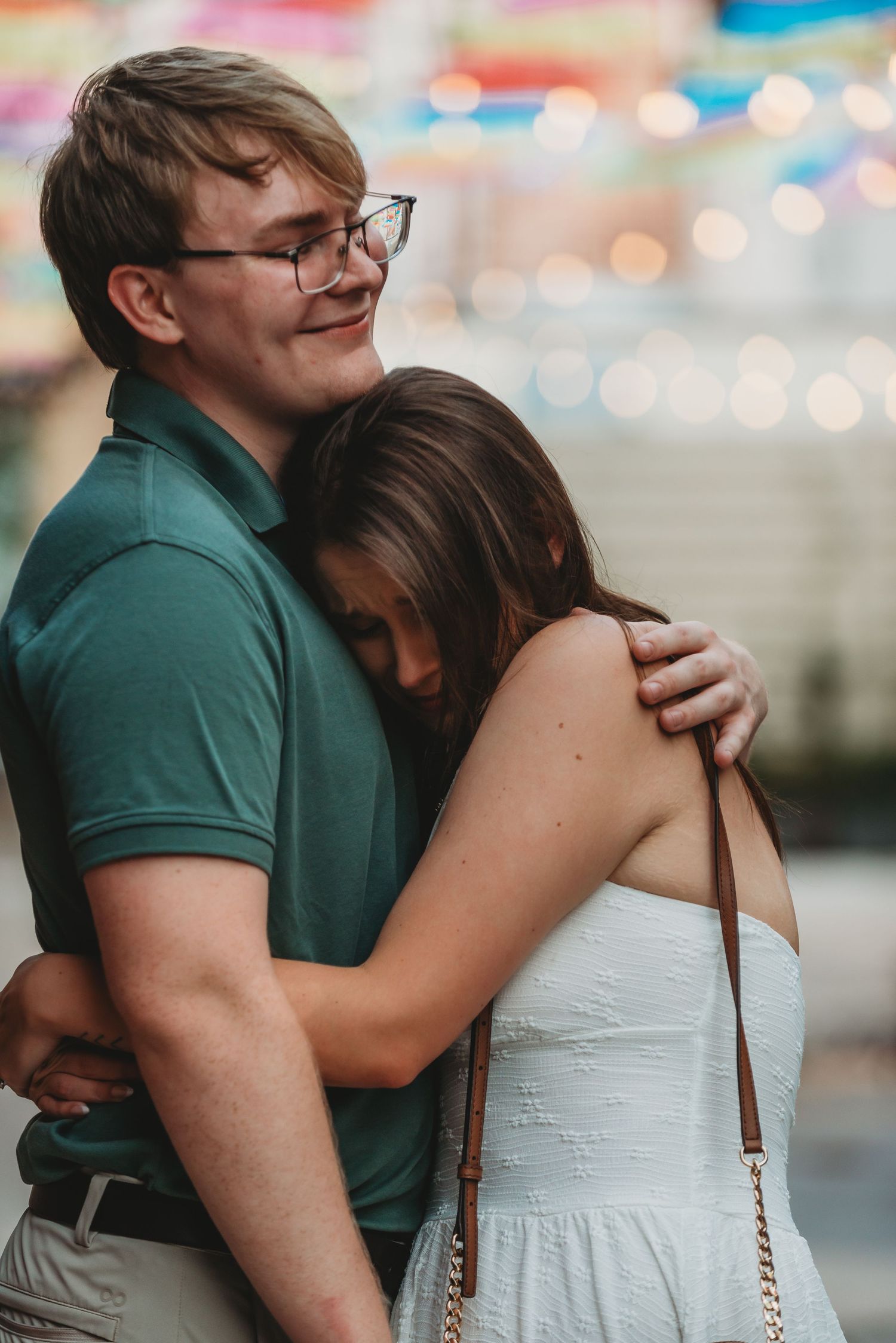 Couple embracing at a colorful fair, man in green polo with glasses hugging woman in white dress.