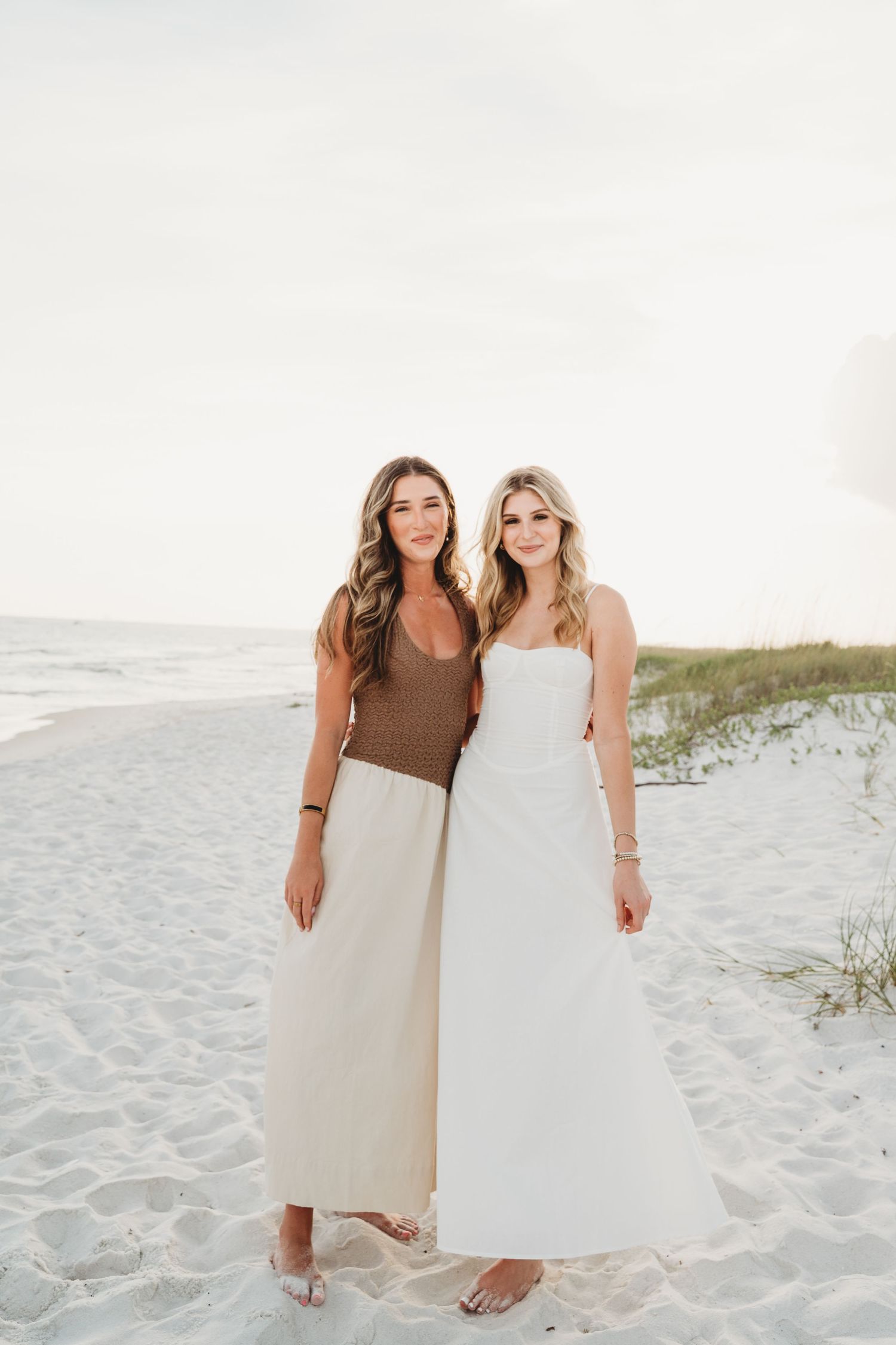 Two young women posing together on a white sand beach, one in brown top and one in white dress.