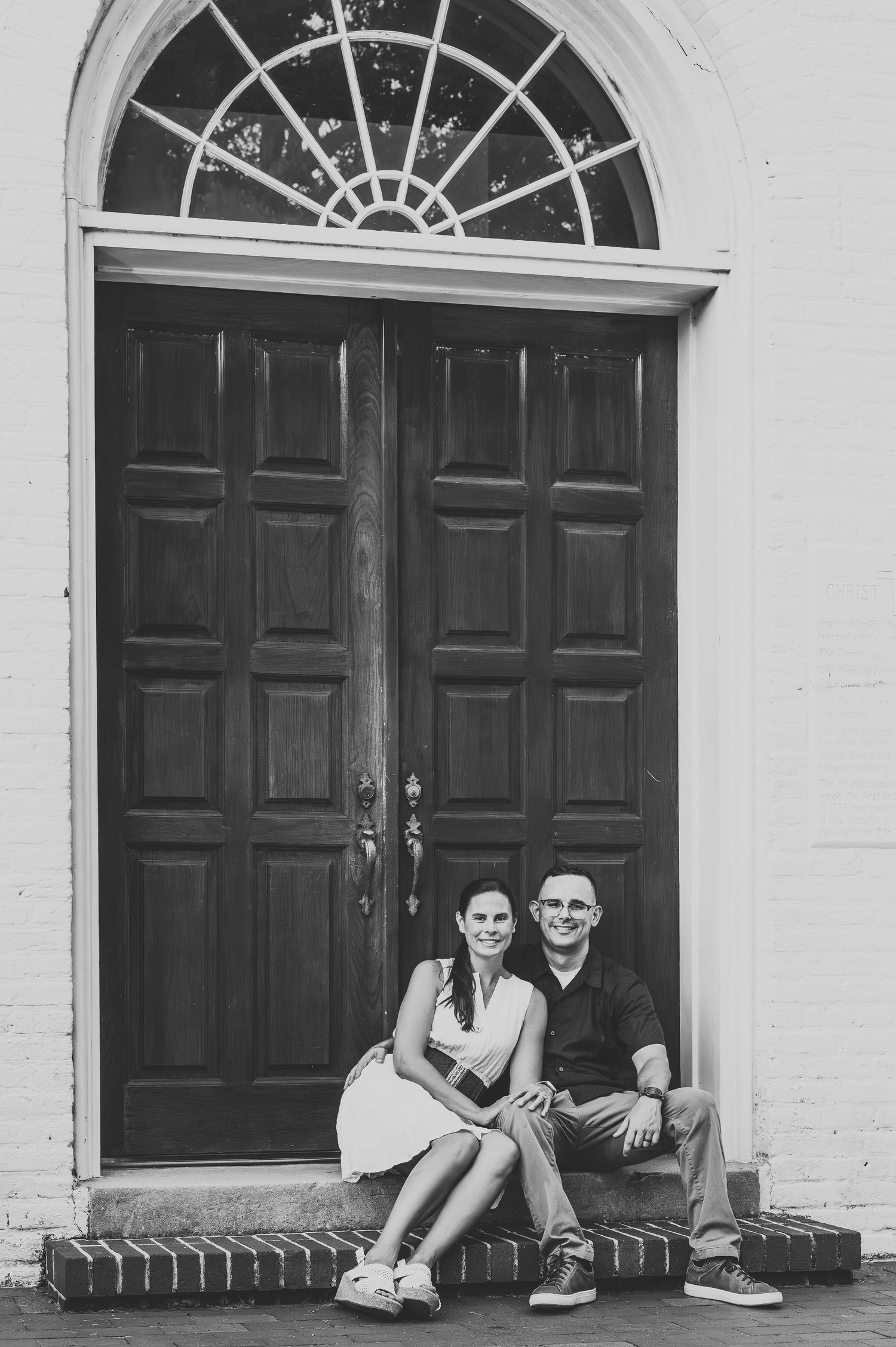 Black and white photo of a couple sitting on brick steps before large wooden double doors.