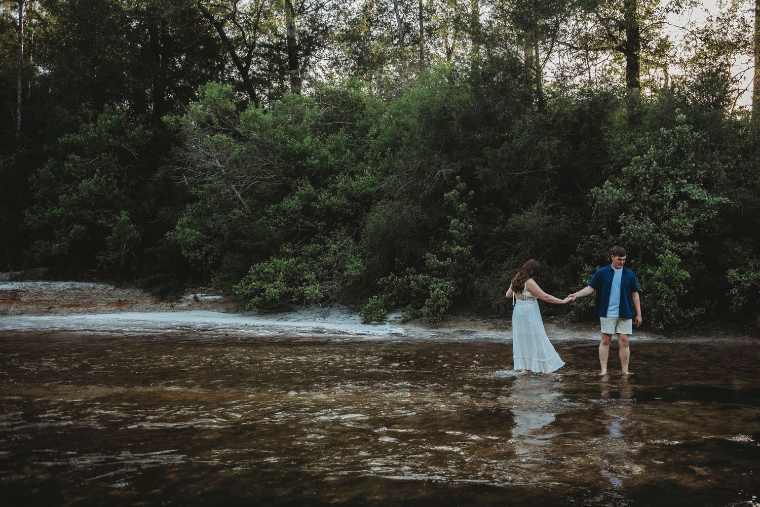 Couple walking hand in hand through a sunlit forest river near a gentle waterfall.