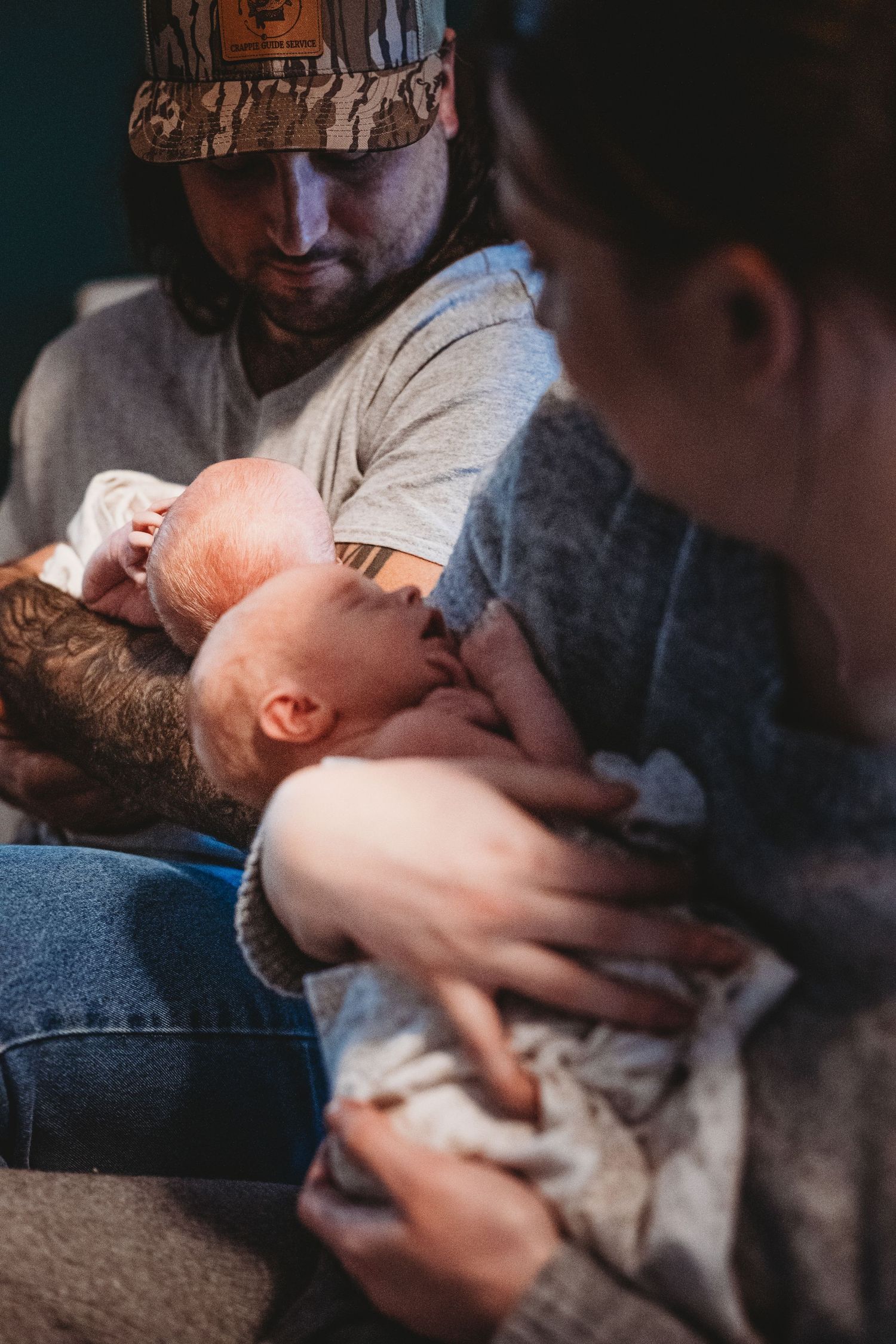 Tattooed father and mother holding twin newborns together in warm indoor setting.