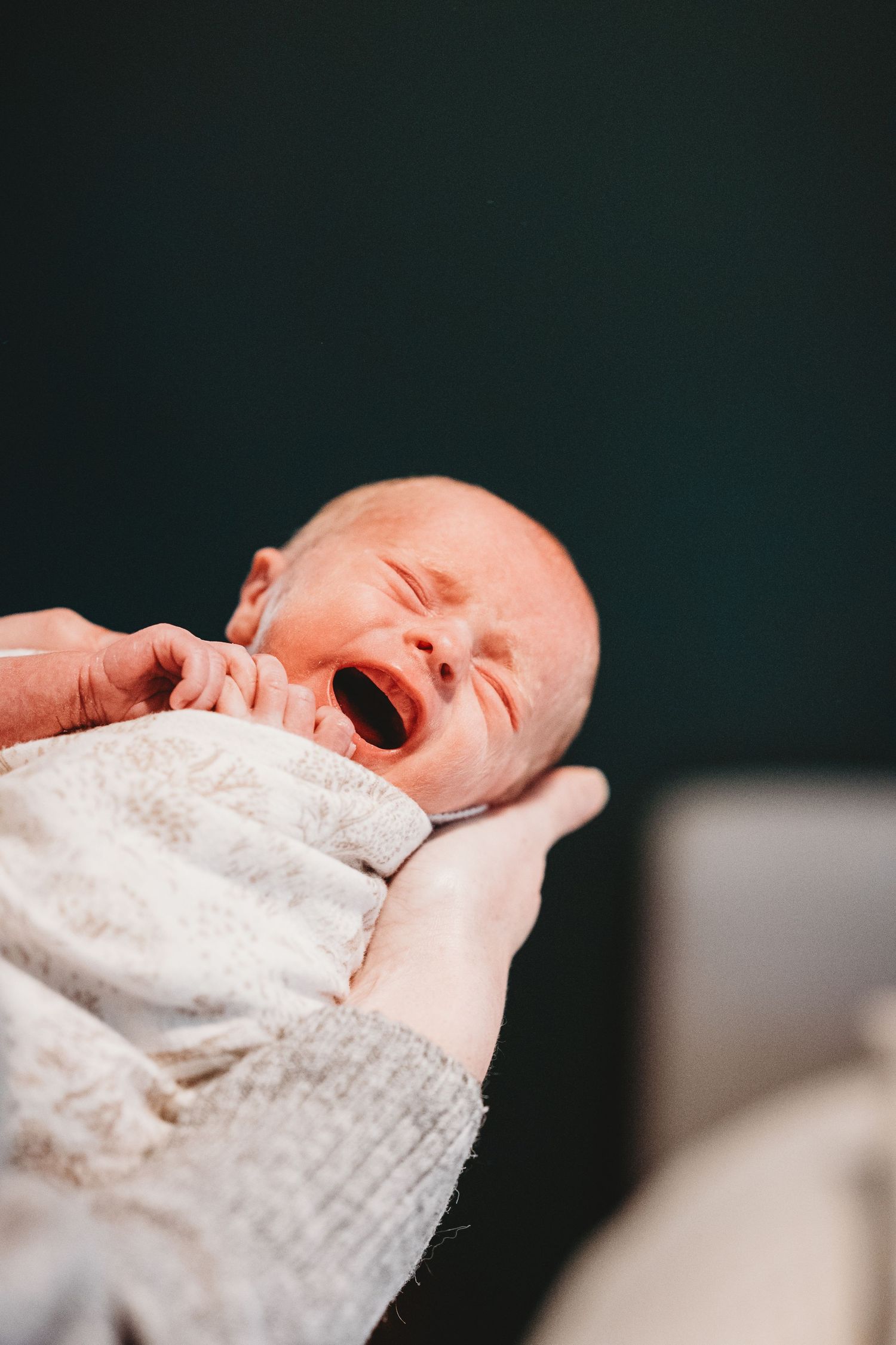 Newborn baby yawning while being cradled in adult hands wrapped in white blanket.