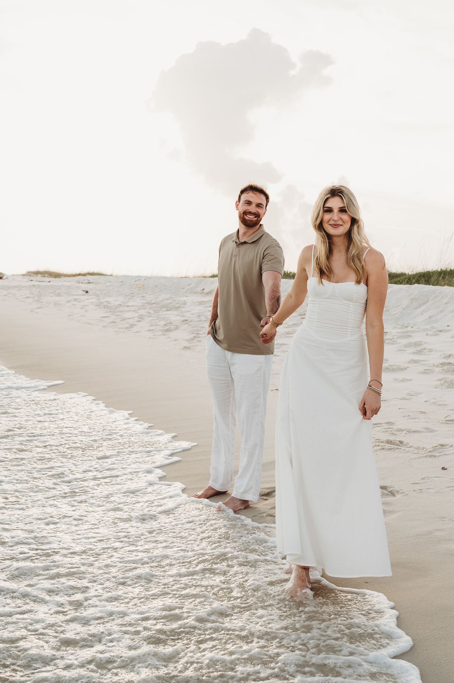 A smiling couple holds hands while walking barefoot along a sandy beach at sunset.