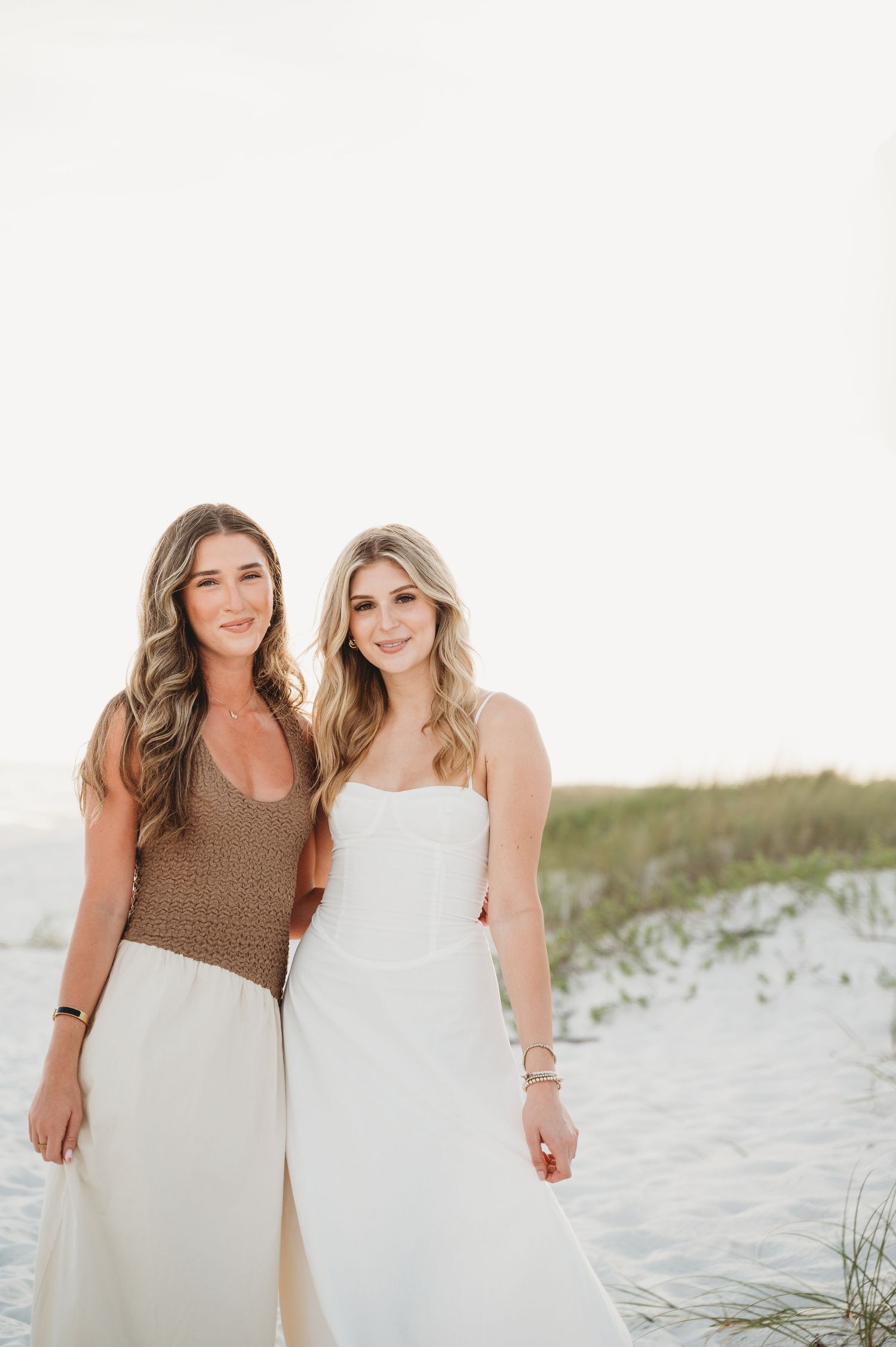 Two smiling women standing together near sand dunes on a beach, one in brown top and one in white dress.
