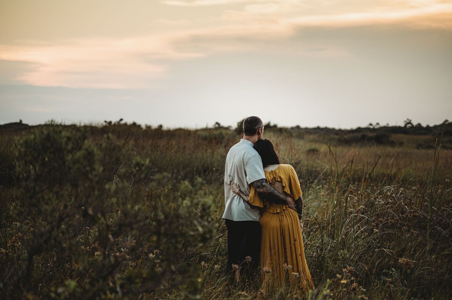 Couple embracing in a golden field of wildflowers at dusk with cloudy sky.