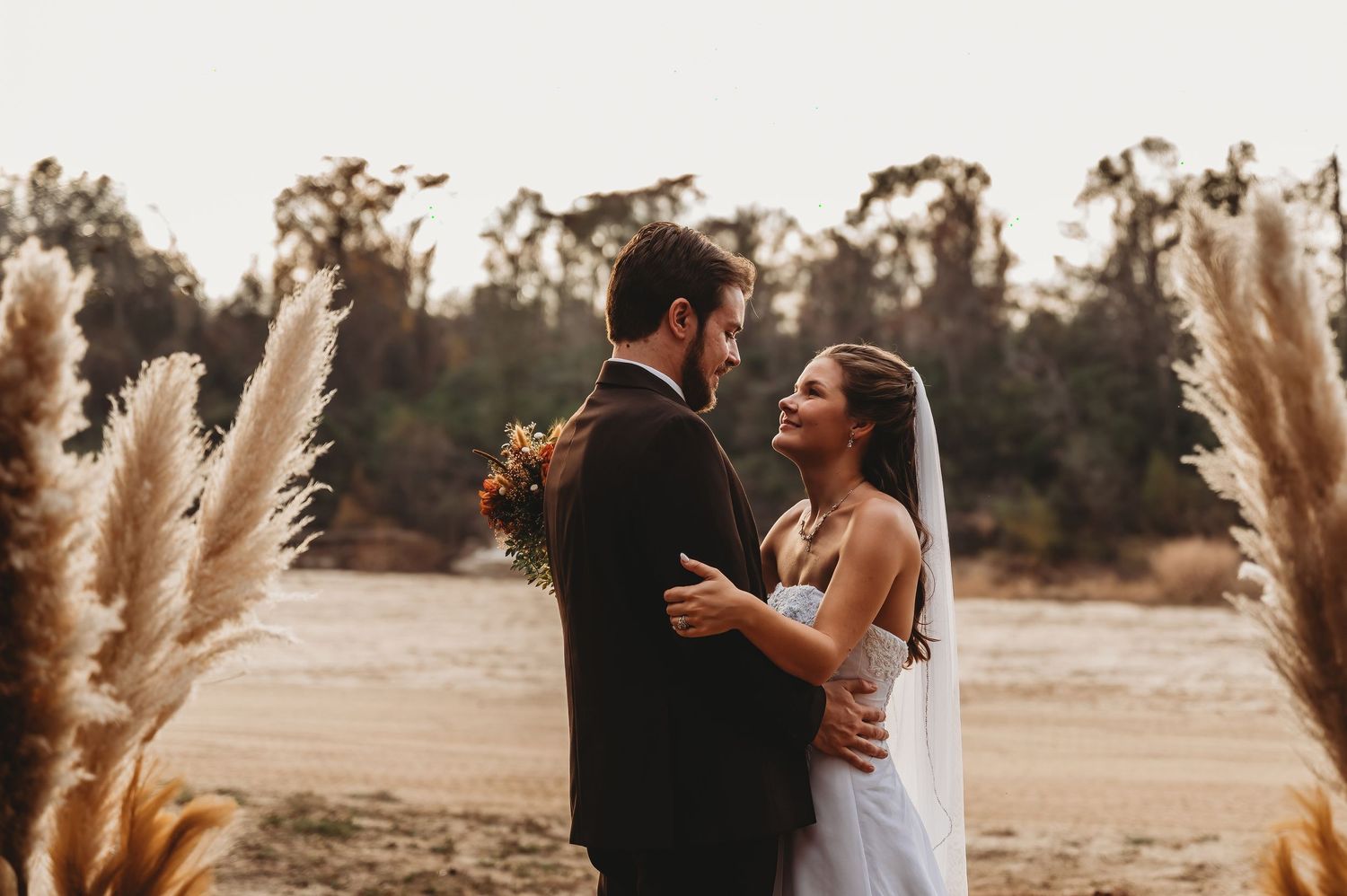 Bride and groom embrace outdoors surrounded by pampas grass at golden hour wedding ceremony.