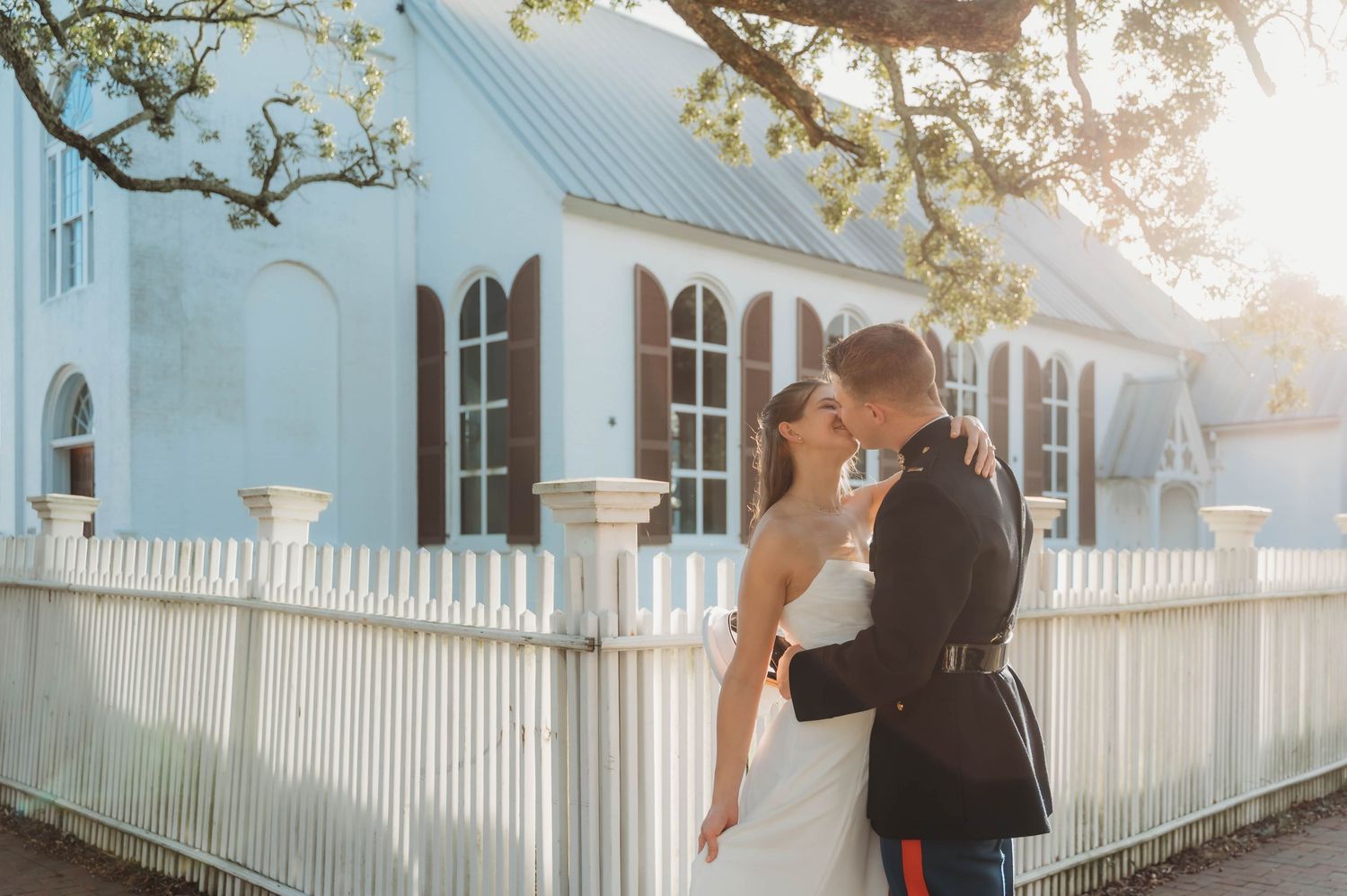 A bride and groom share a romantic kiss outside a charming blue church with a white picket fence.
