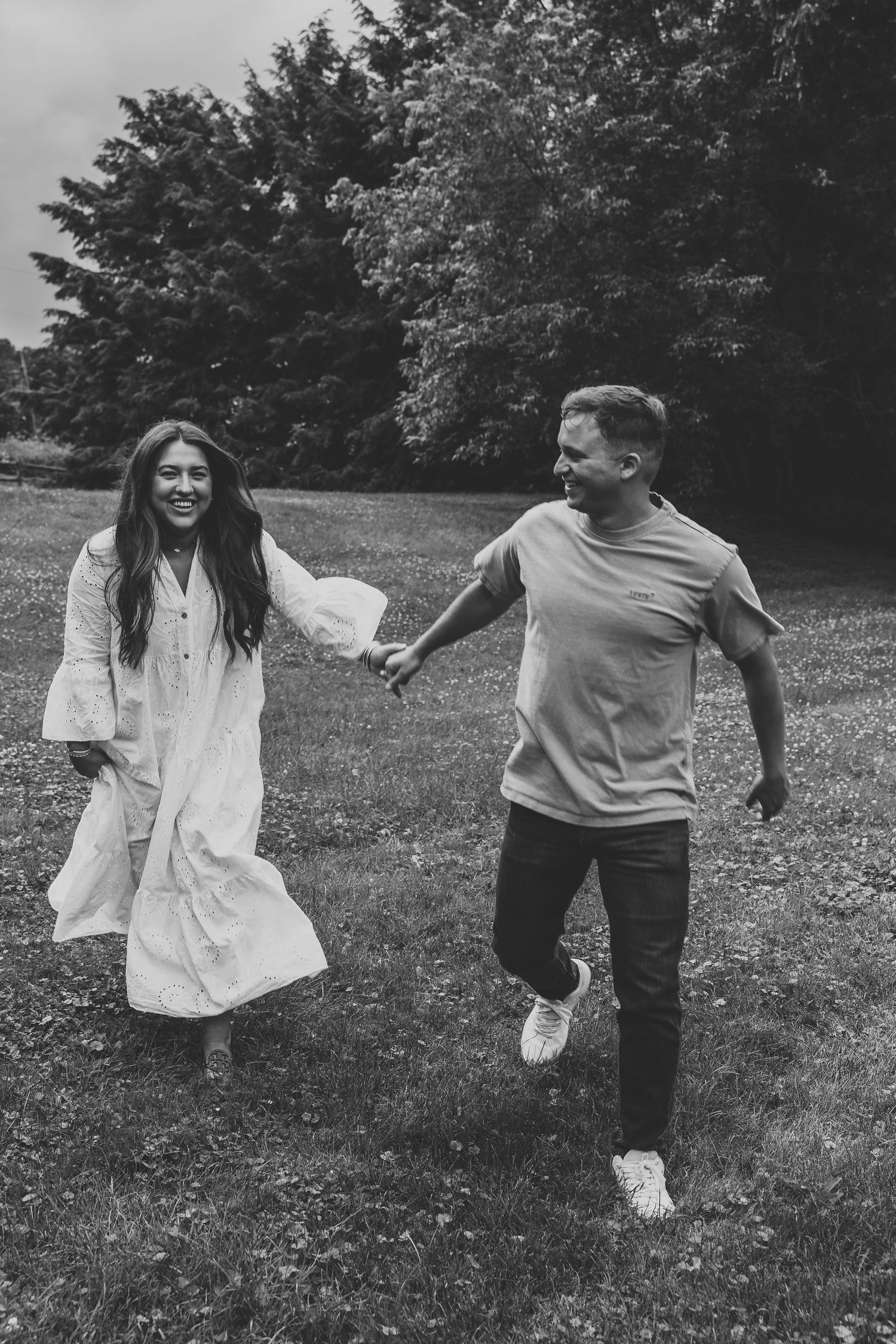 Black and white photo of a joyful couple holding hands and running through a lush green park together.