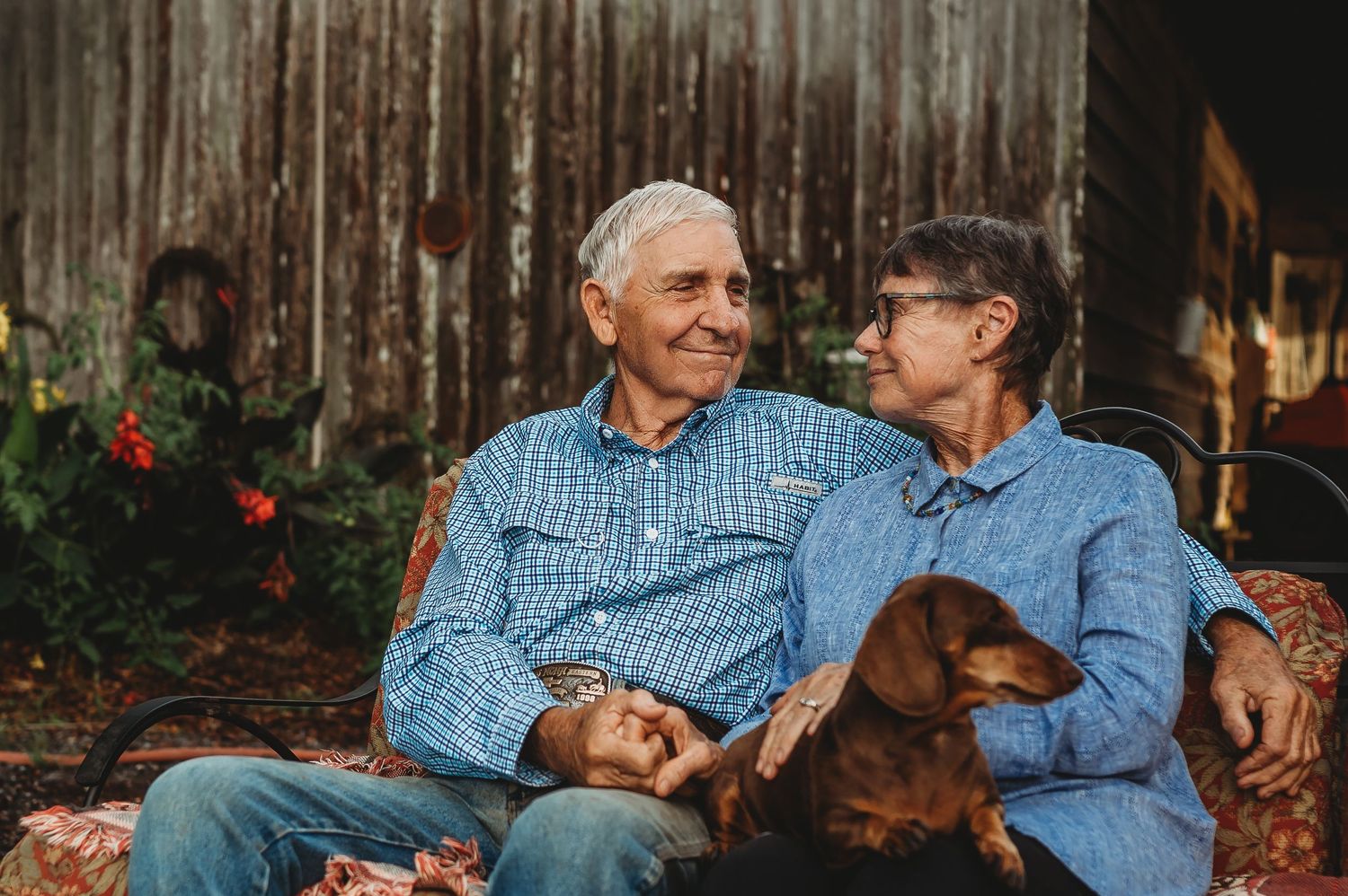 Elderly couple smiling at each other on a bench with a dachshund dog, seated outdoors near a rustic barn.