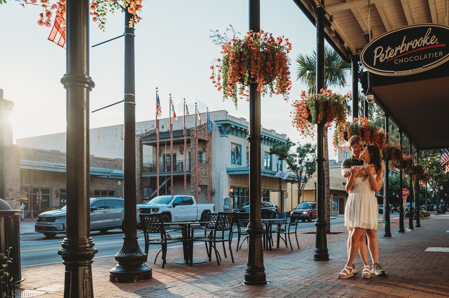Woman in white dress strolling past Peterbrooke Chocolatier on a sunny brick-lined street with hanging flower baskets.