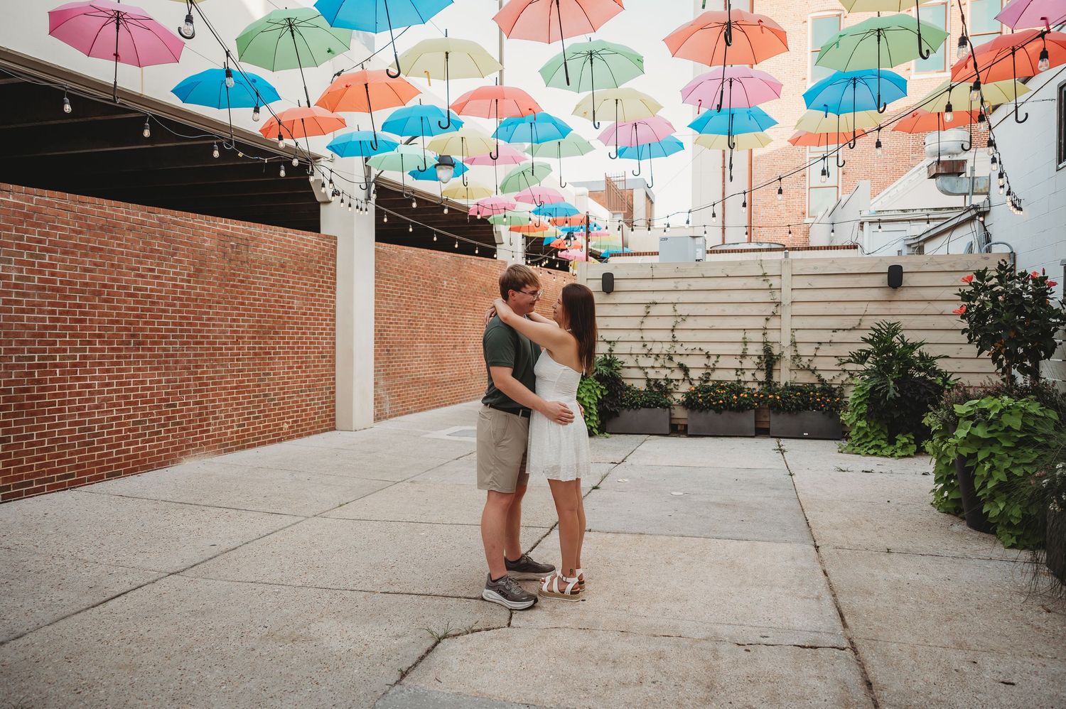 Couple embracing under colorful hanging umbrellas in an outdoor courtyard with string lights and brick wall backdrop.