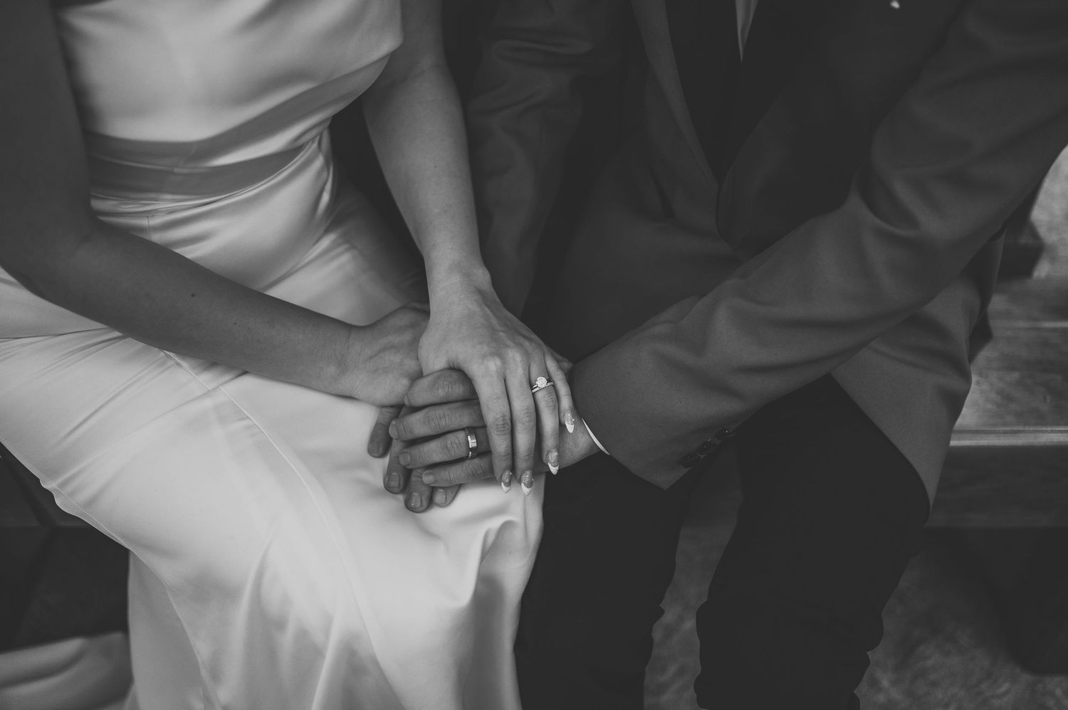 Black and white photo of bride and groom holding hands, wedding rings visible, romantic close-up shot.