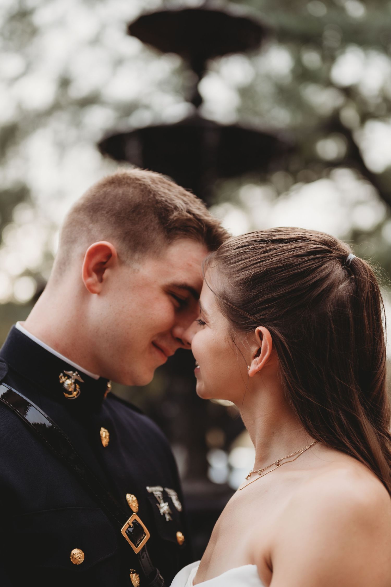 A Marine in dress uniform and bride share an intimate forehead touch near a fountain on their wedding day.