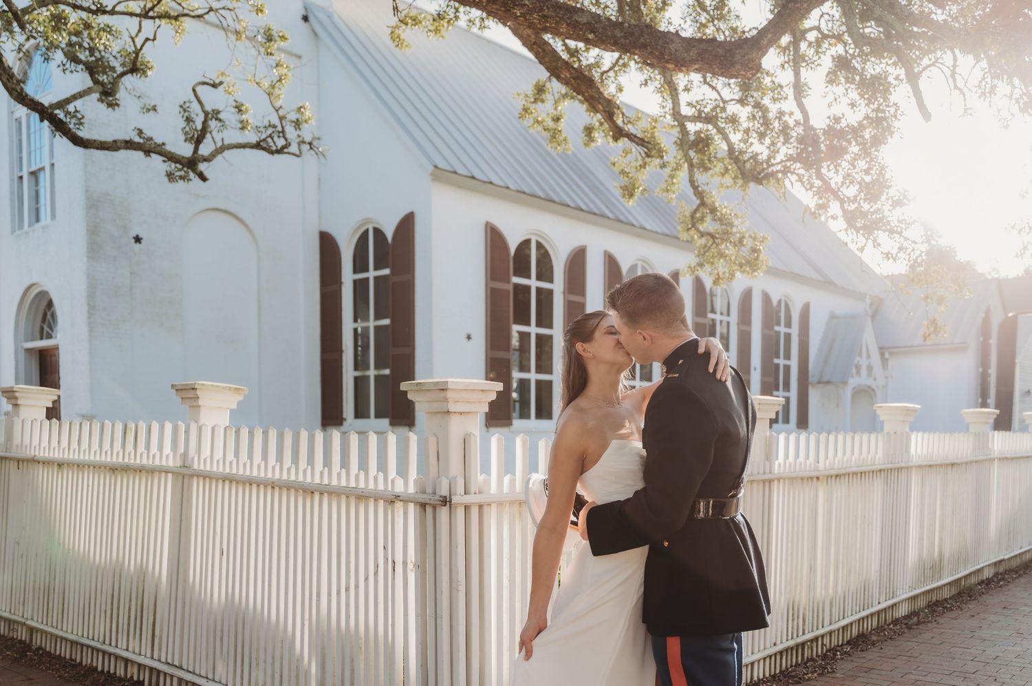 A couple shares a romantic kiss near a white picket fence outside a charming blue church on their wedding day.
